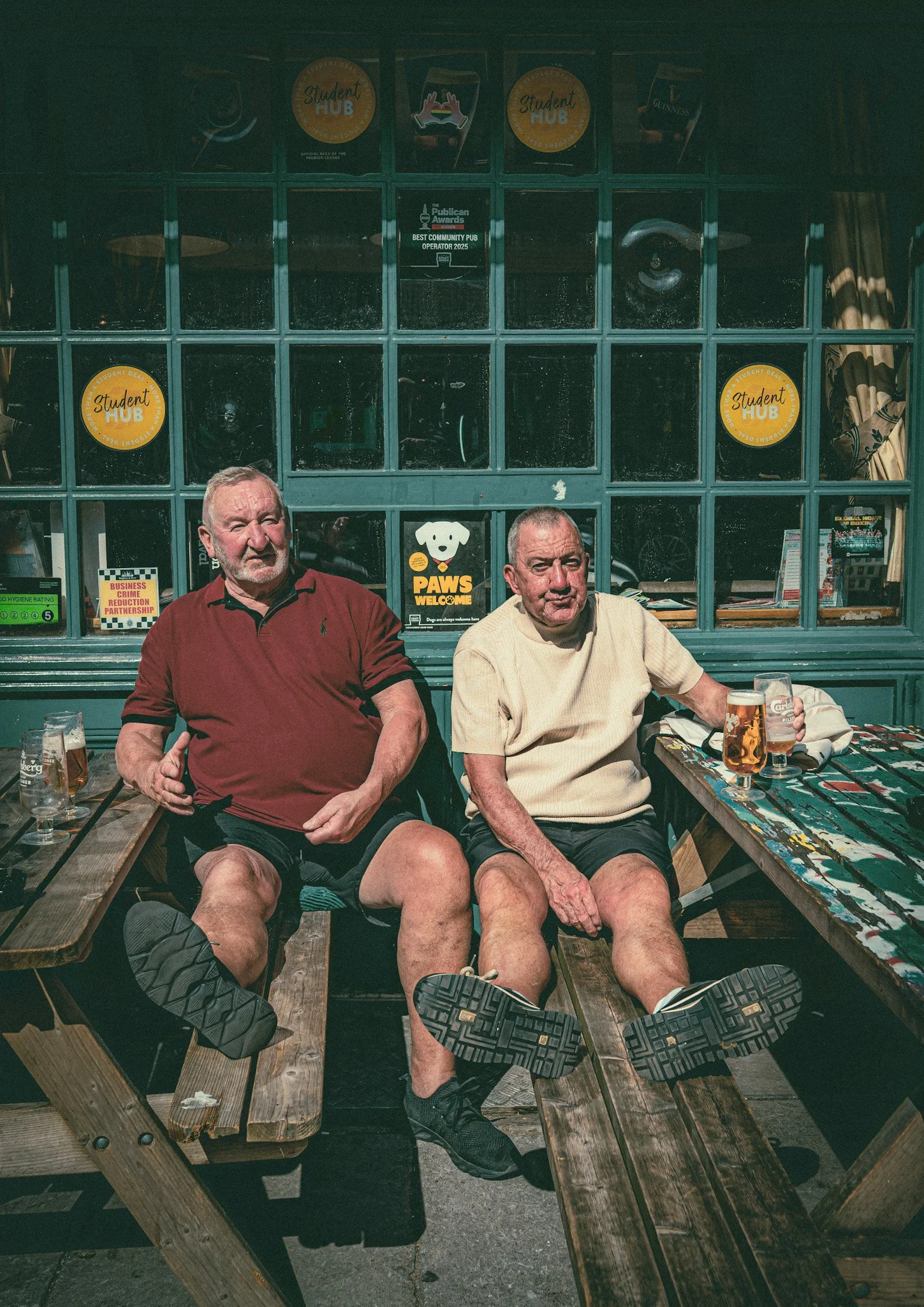 Two elderly men sitting on a wooden bench outdoors, with their legs extended and feet up. They are drinking beer, and the background features a rustic building with windows, signs, and some curtains.