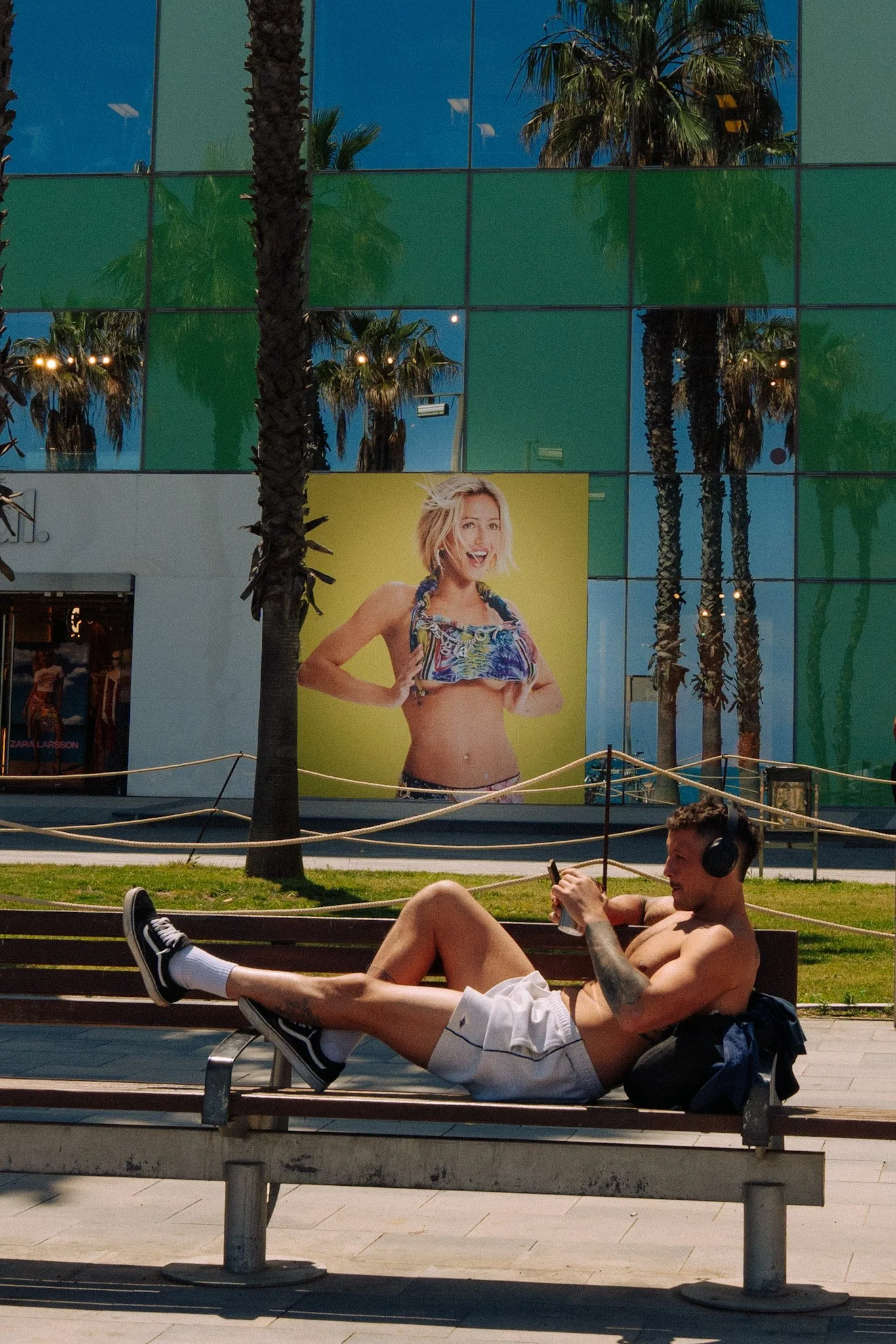 A shirtless man with tattoos and white shorts lying on a park bench, wearing sneakers and headphones, holding a drink. Behind him is a lamppost, green grass, and a large building with trees and colorful windows. A large advertisement with a smiling w