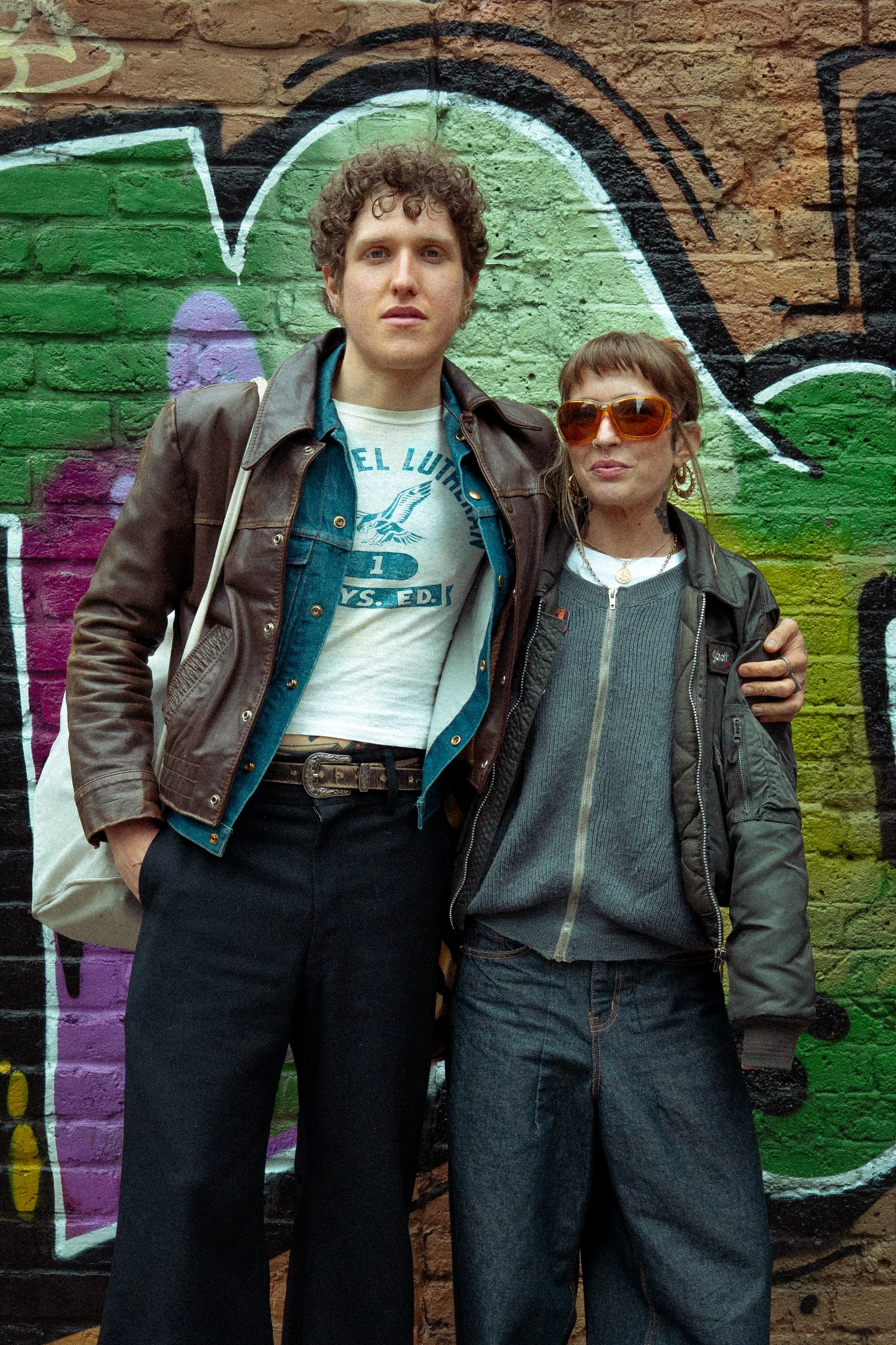 Two people standing in front of a colorful graffiti wall. The person on the left has short curly hair, fair skin, and is wearing a brown jacket over a denim shirt and a graphic T-shirt. The person on the right has short brown hair, tan skin, orange s