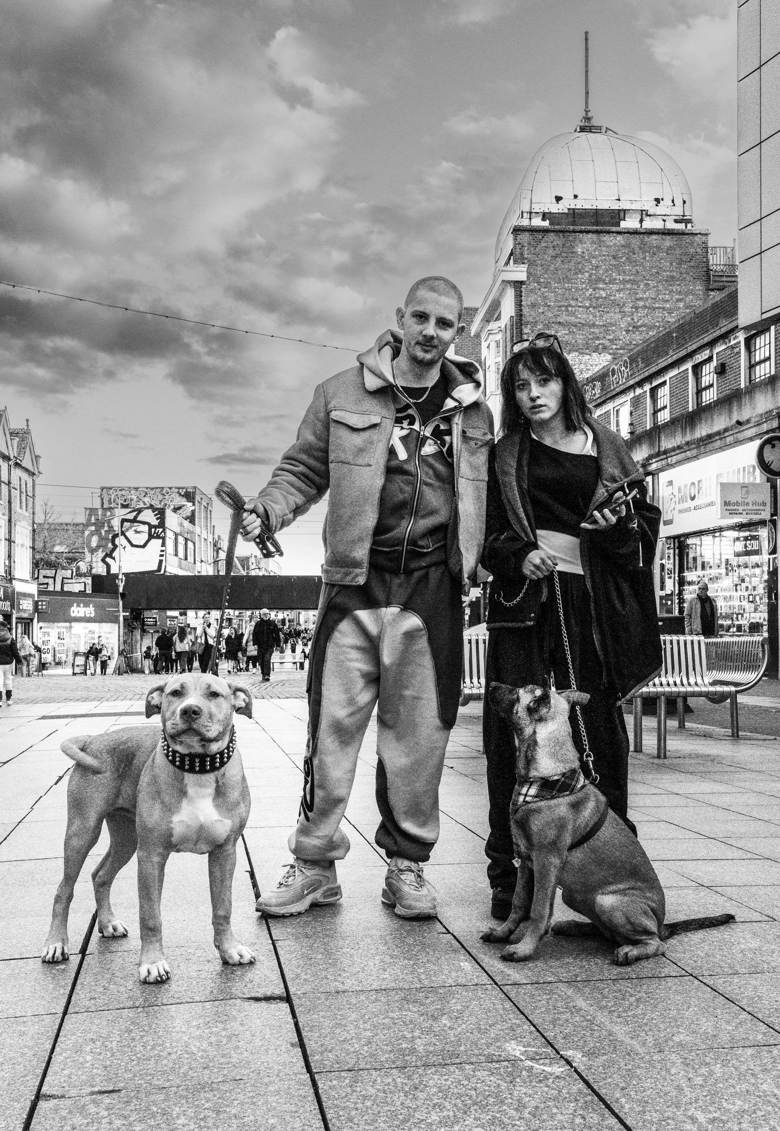 A black and white photo of two people with their dogs in an urban setting. The man is holding a leash attached to a large dog with a spiked collar, and the woman is holding a leash attached to a smaller dog with a plaid coat. The man is wearing a hoo
