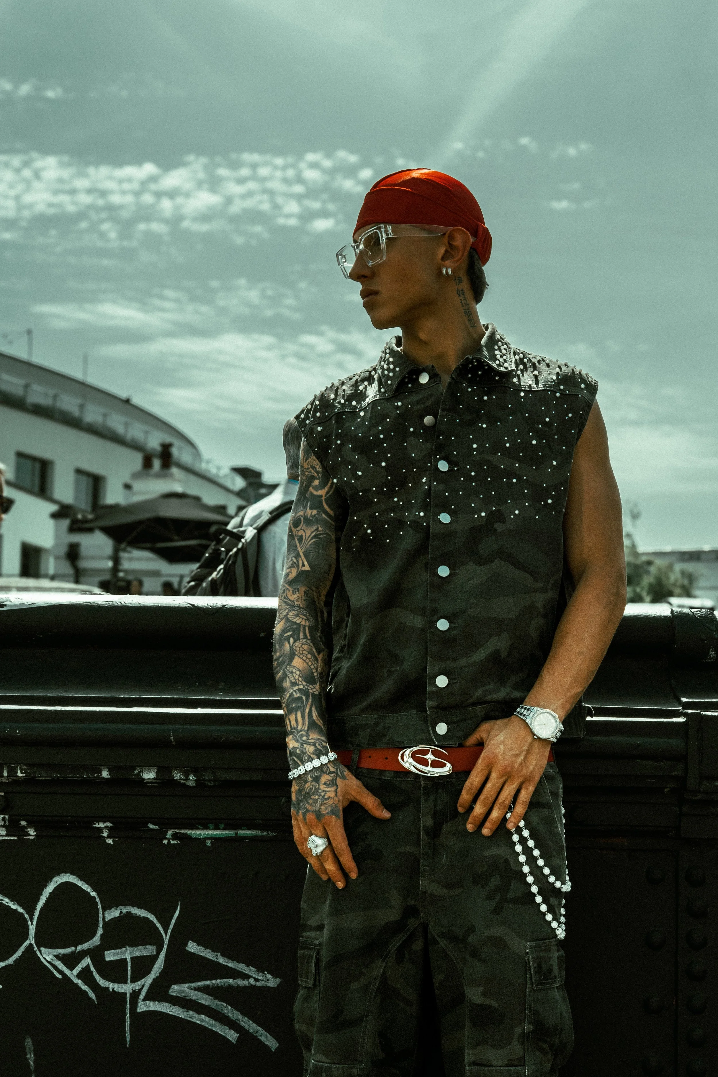 A man with tattoos wearing a red bandana, clear glasses, a sleeveless black vest with white studs, a red belt with a silver buckle, and dark camouflage pants, standing outdoors against a black surface with graffiti, under a cloudy sky.