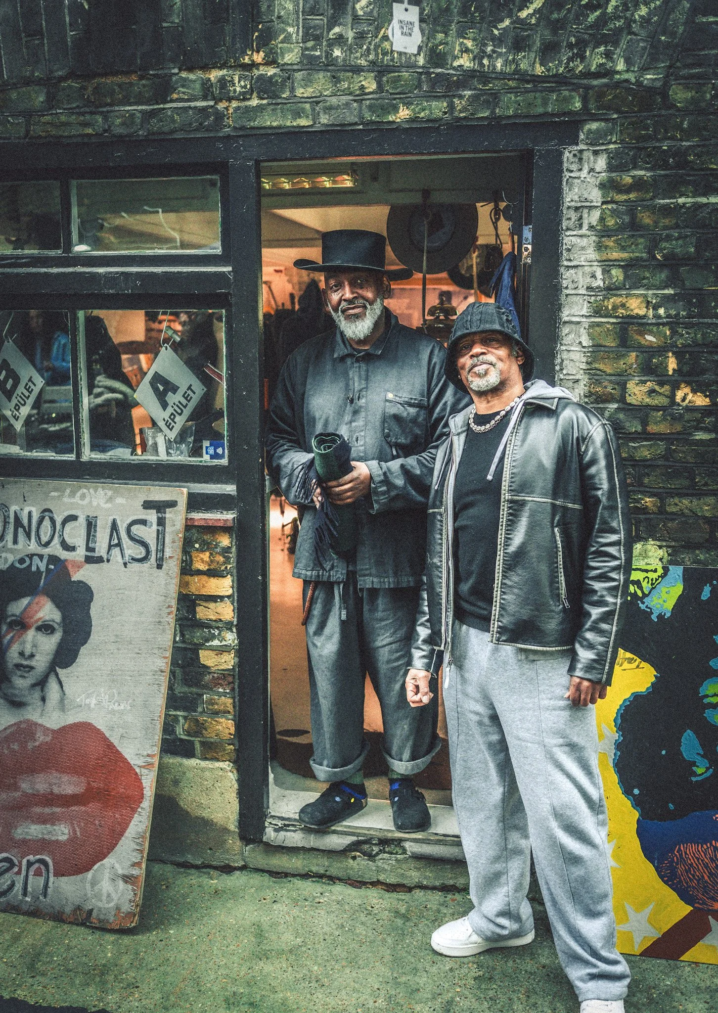 Two men standing in front of a brick building entrance, one wearing a black cowboy hat and leather jacket, the other wearing a bucket hat and grey sweatpants, in an urban setting with posters and artwork visible.