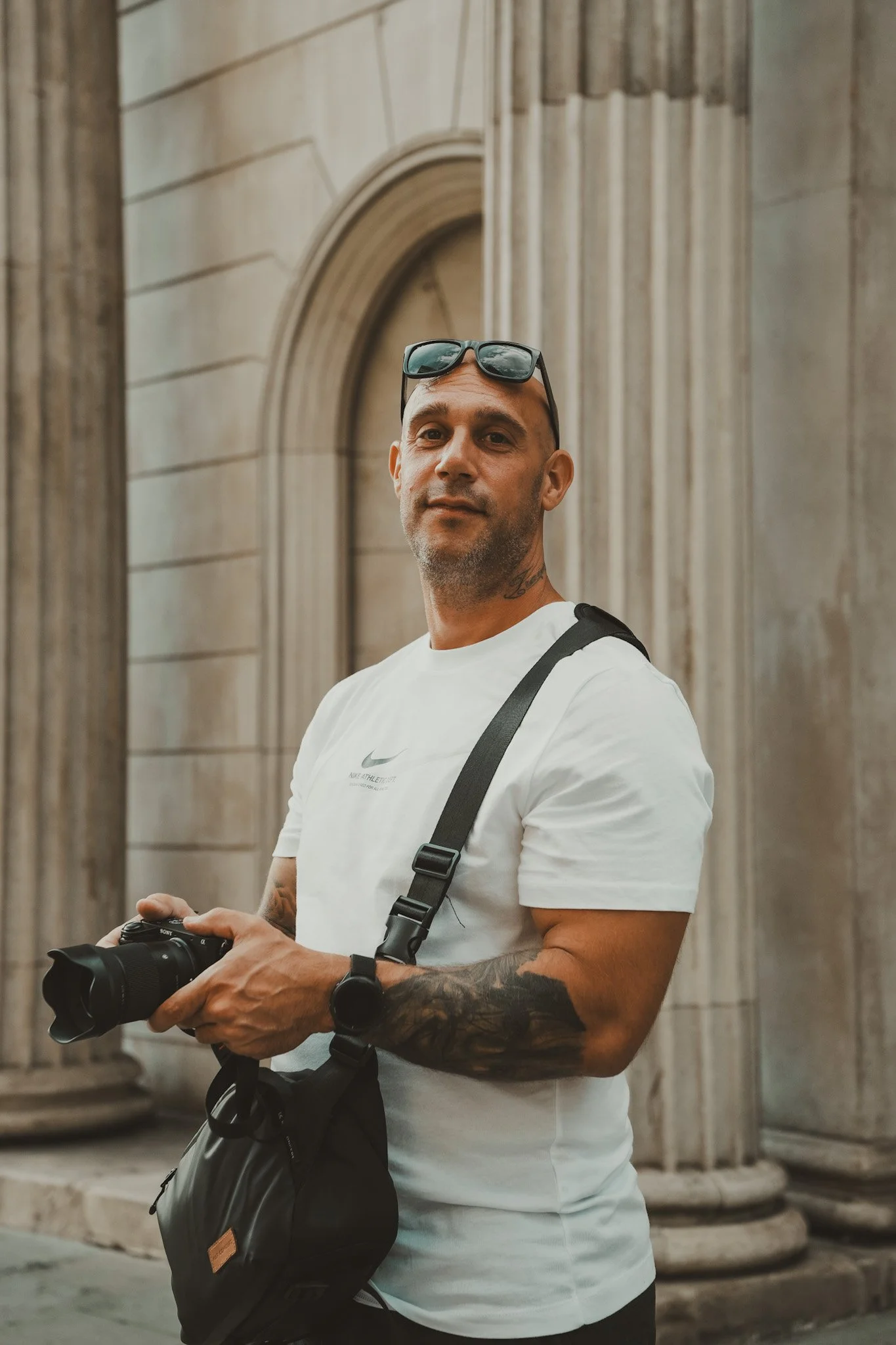 A man with sunglasses on his head, wearing a white Nike T-shirt, holding a camera, and carrying a black bag stands in front of stone columns and arches.