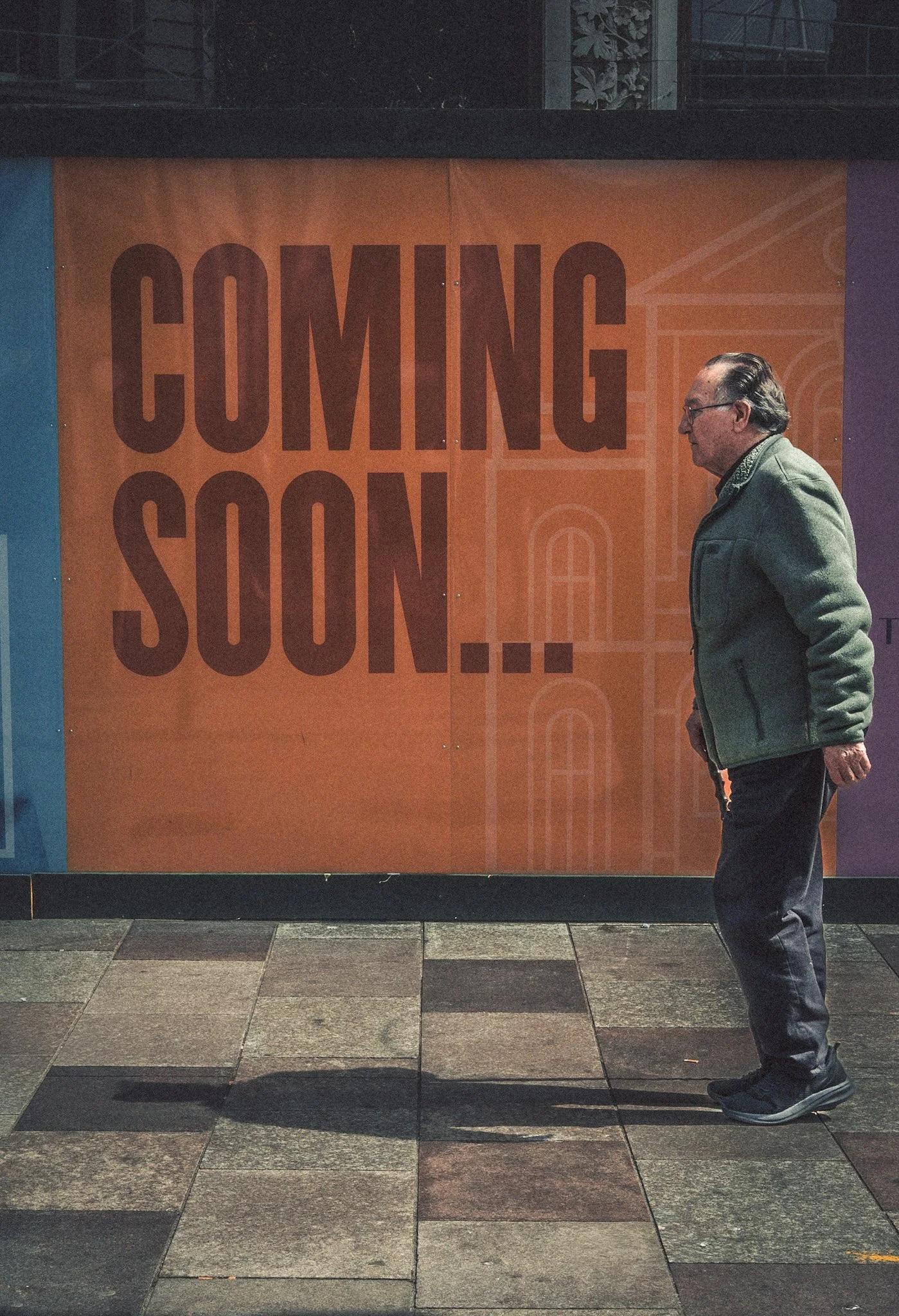 A man in a green jacket and dark pants walking past a large orange poster with the text 'COMING SOON...' displayed on a city sidewalk at night.