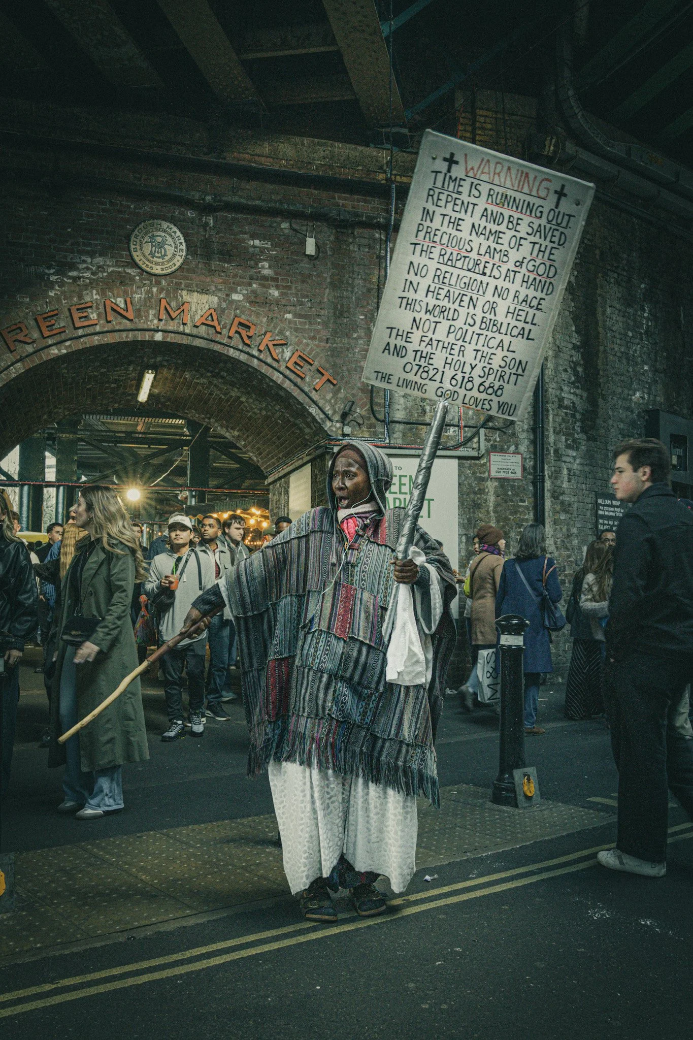 A person dressed in traditional attire holding a religious sign with biblical text, standing among a crowd in an urban setting under a bridge or archway.