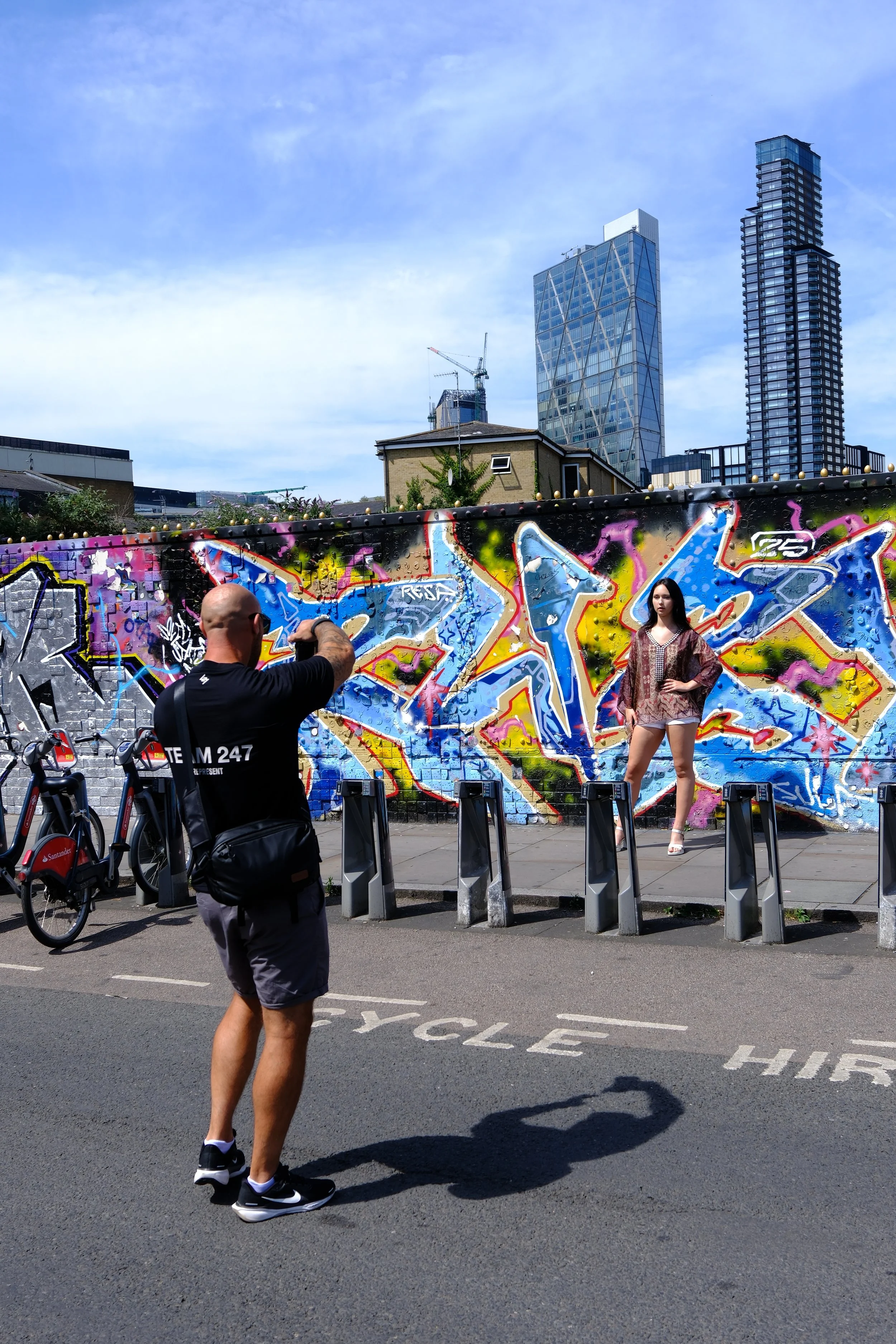 A man taking a photo of a woman posing in front of a colorful graffiti wall in an urban setting during daytime. The man is wearing black shorts and a black T-shirt, and the woman is dressed in a patterned blouse and shorts. Behind the wall, modern skyscrapers can be seen, with a clear blue sky overhead.