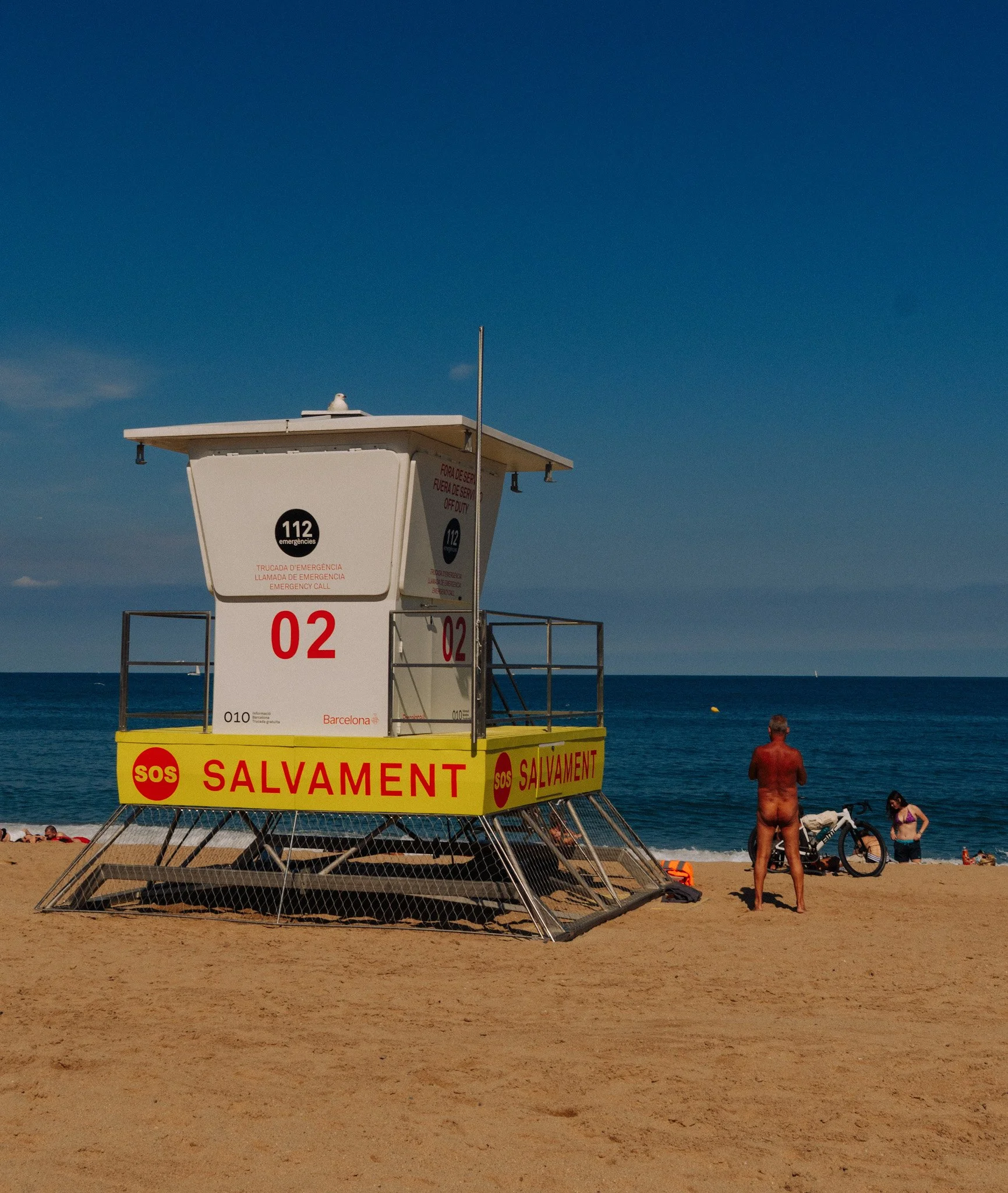 A lifeguard tower on a beach with a seagull perched on top, and a person in a bathing suit sitting nearby with a bicycle and some belongings on the sand, under a clear blue sky.