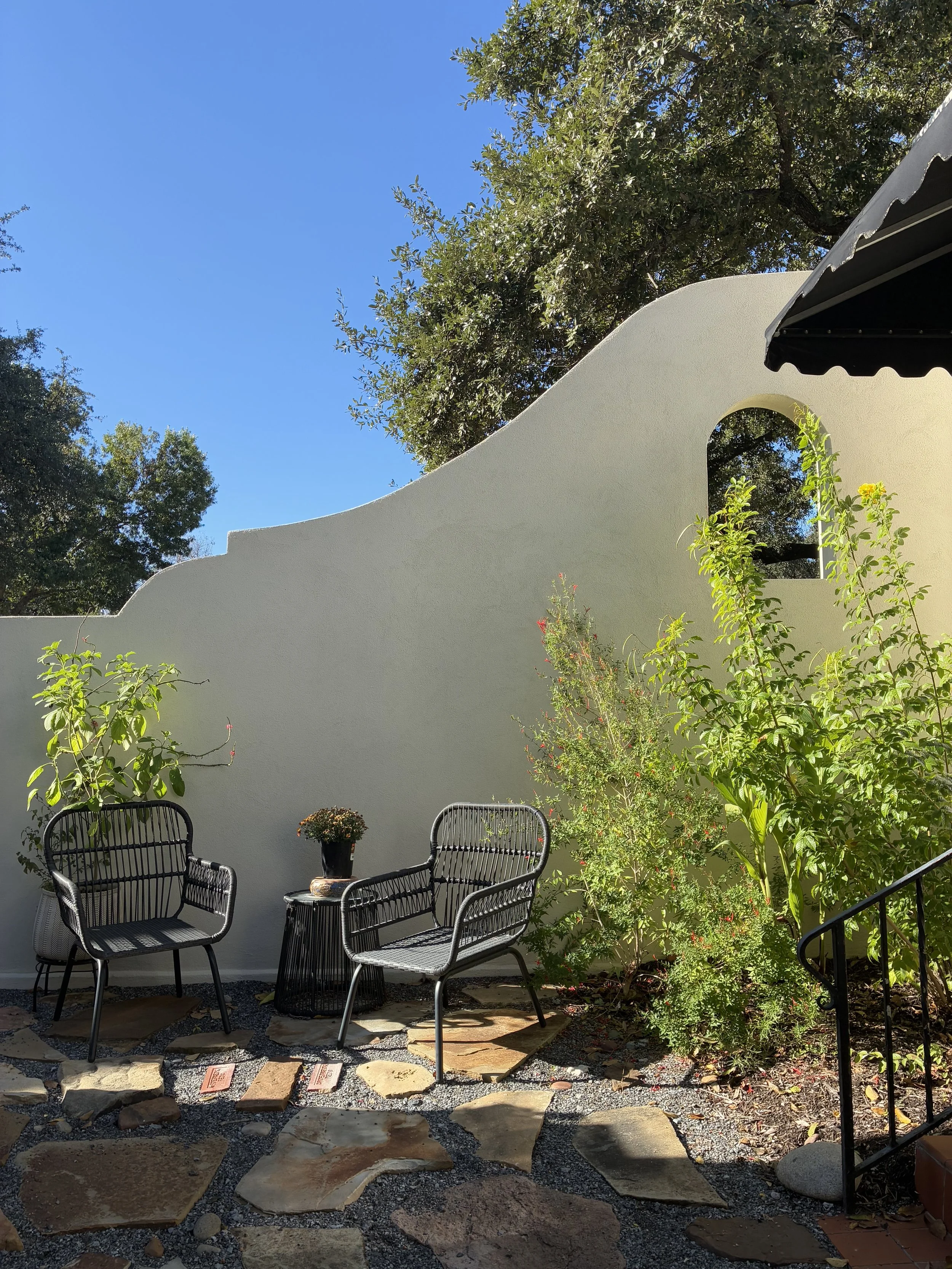 A cozy outdoor patio with black wicker chairs and a small table with a potted plant. The patio is decorated with various green plants and flowers. There's a tall white wall with an arched opening, and the sky is clear and blue with trees in the background.
