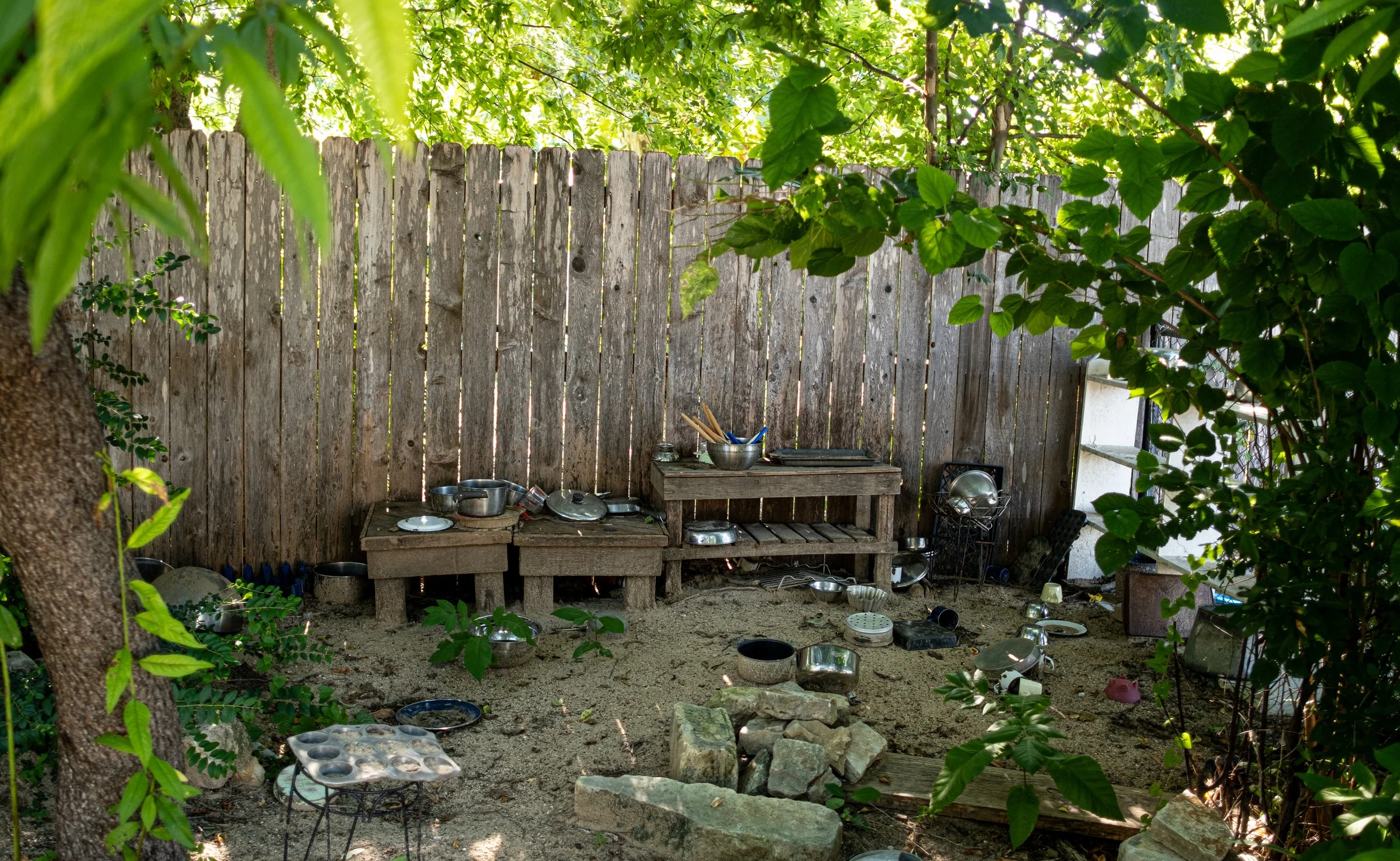 An outdoor area with a wooden fence, cluttered with various metal bowls and dishes, surrounded by greenery and trees, with sunlight filtering through the leaves.