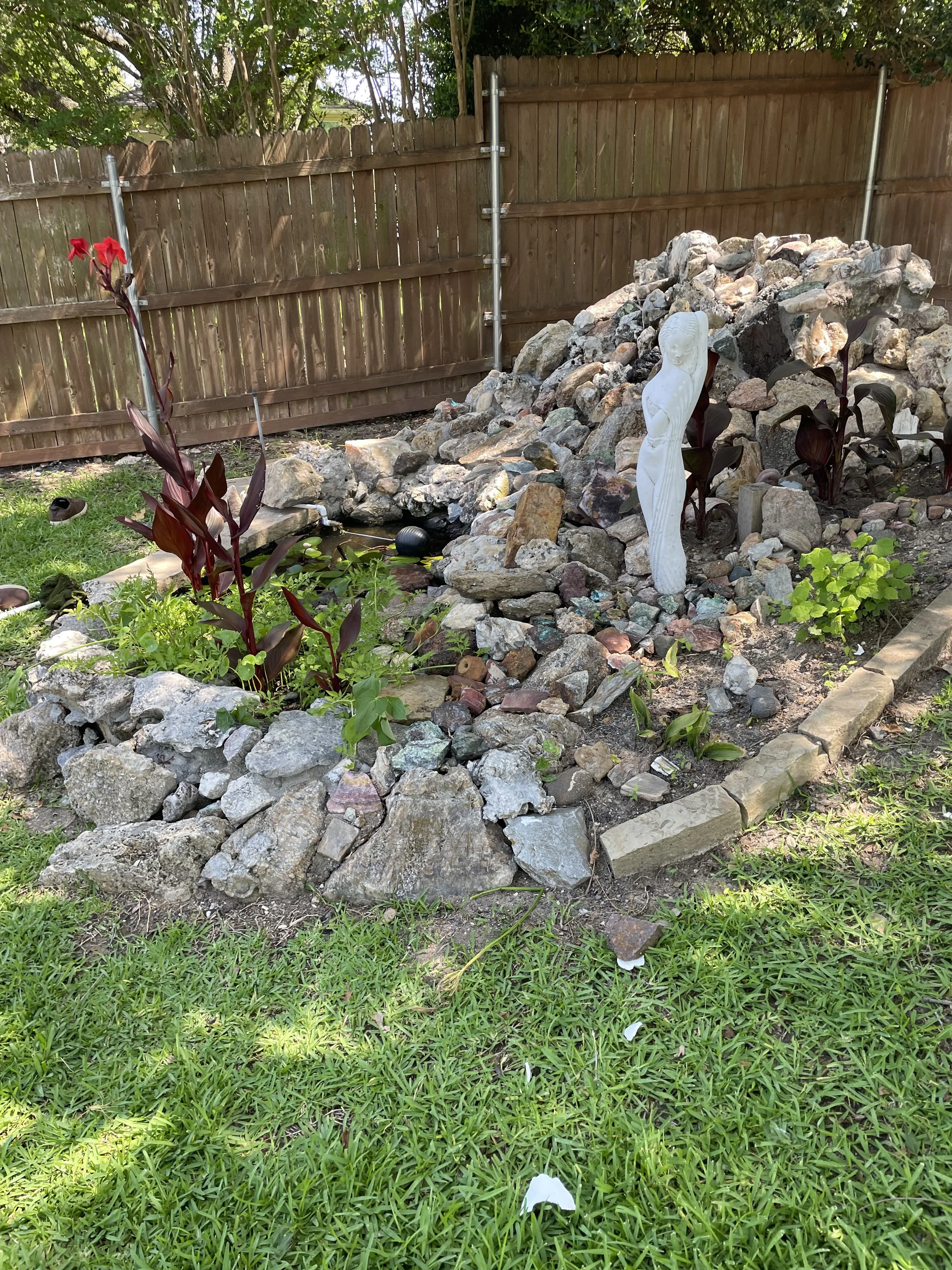 A backyard garden with a spider-man decorated rock water fountain, surrounded by green grass, various plants, rocks, and a wooden fence in the background.