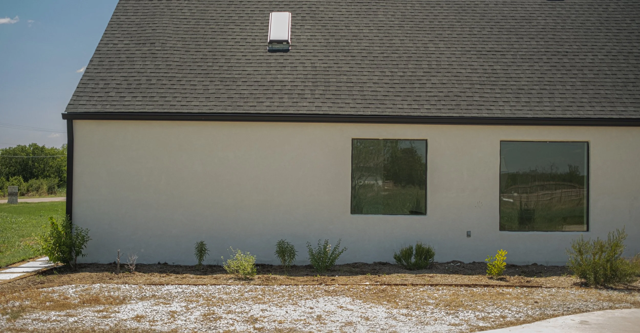 A modern house with white exterior walls, two large windows, a dark shingle roof, and a skylight, with a garden featuring small plants and flowers in the foreground.
