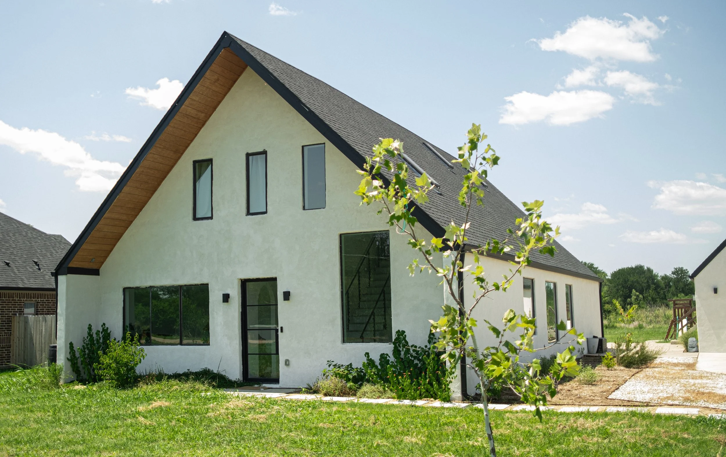 Modern house with a steep gable roof, multiple tall narrow windows, and a black front door, surrounded by a lawn and small trees on a sunny day with a partly cloudy sky.