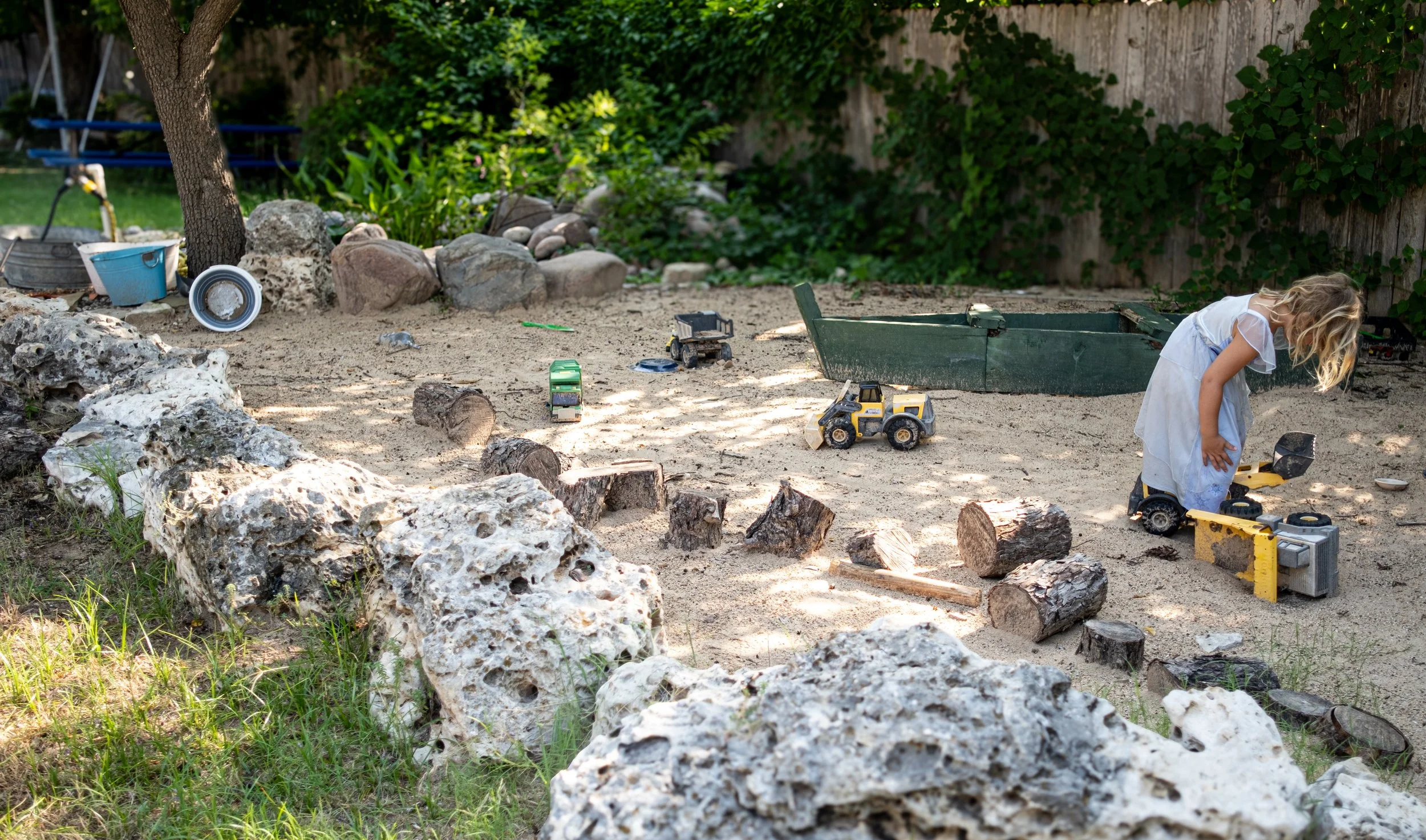 A young girl with blonde hair in a white dress is playing in a sandbox with toy construction trucks. The sandbox is surrounded by large rocks and logs, with green plants and ivy-covered fence in the background.