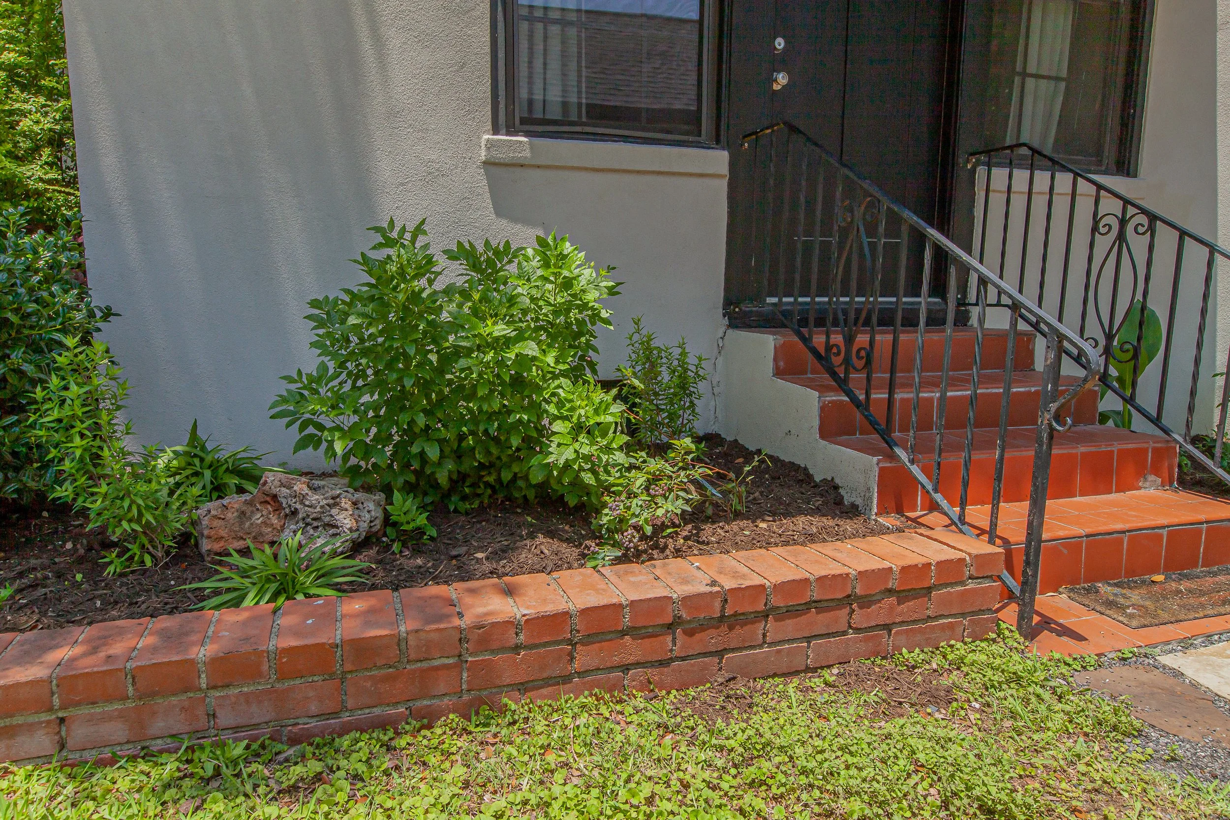 A front porch with four steps leading up to a black door with a glass window and a security screen. The porch has a black metal railing and red tiles. There are green bushes and plants around the base of the stairs, and a white wall to the left with 