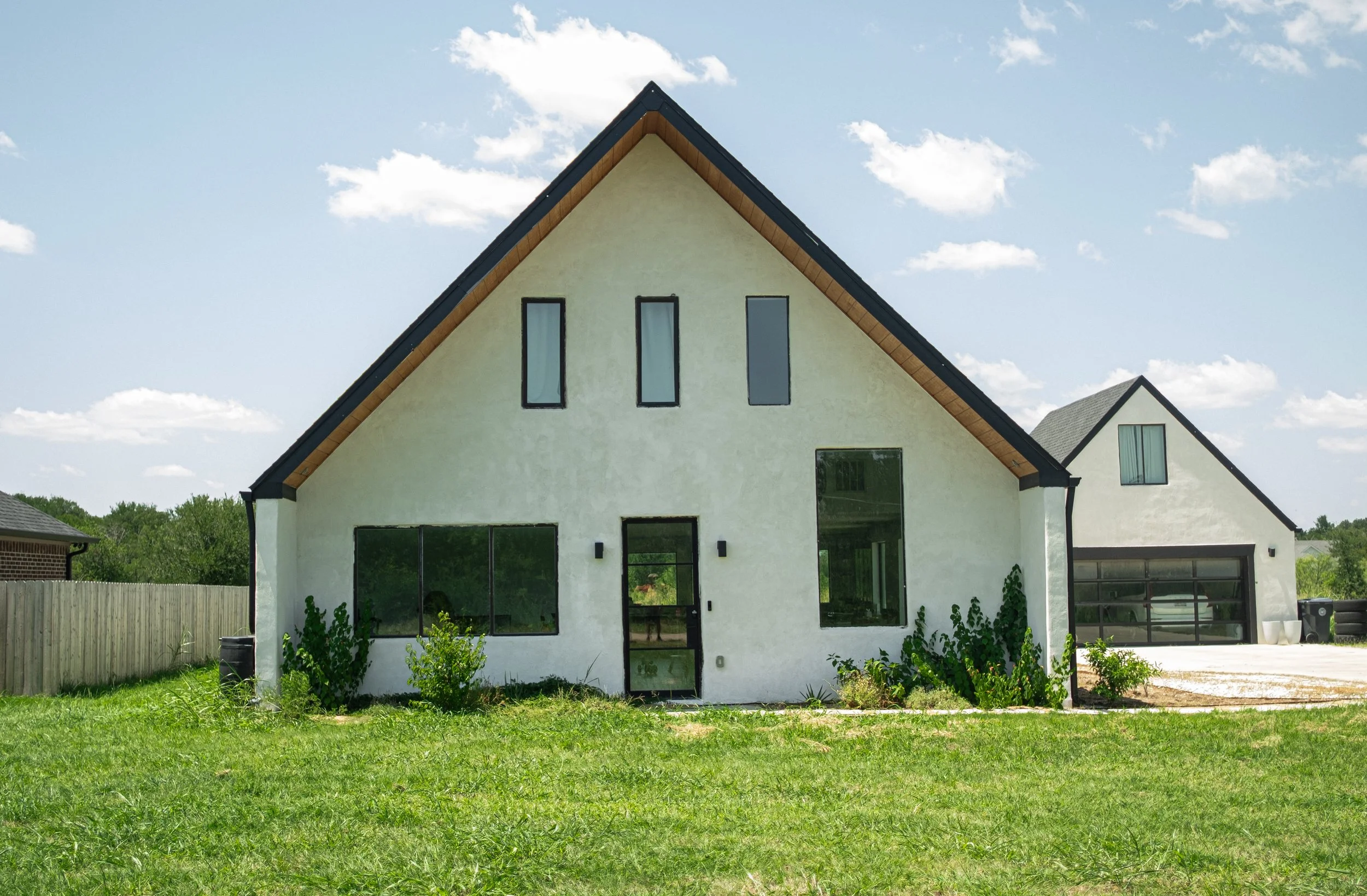 Modern white house with large windows, black trim, a steep gabled roof, and a fenced backyard, under a partly cloudy sky.
