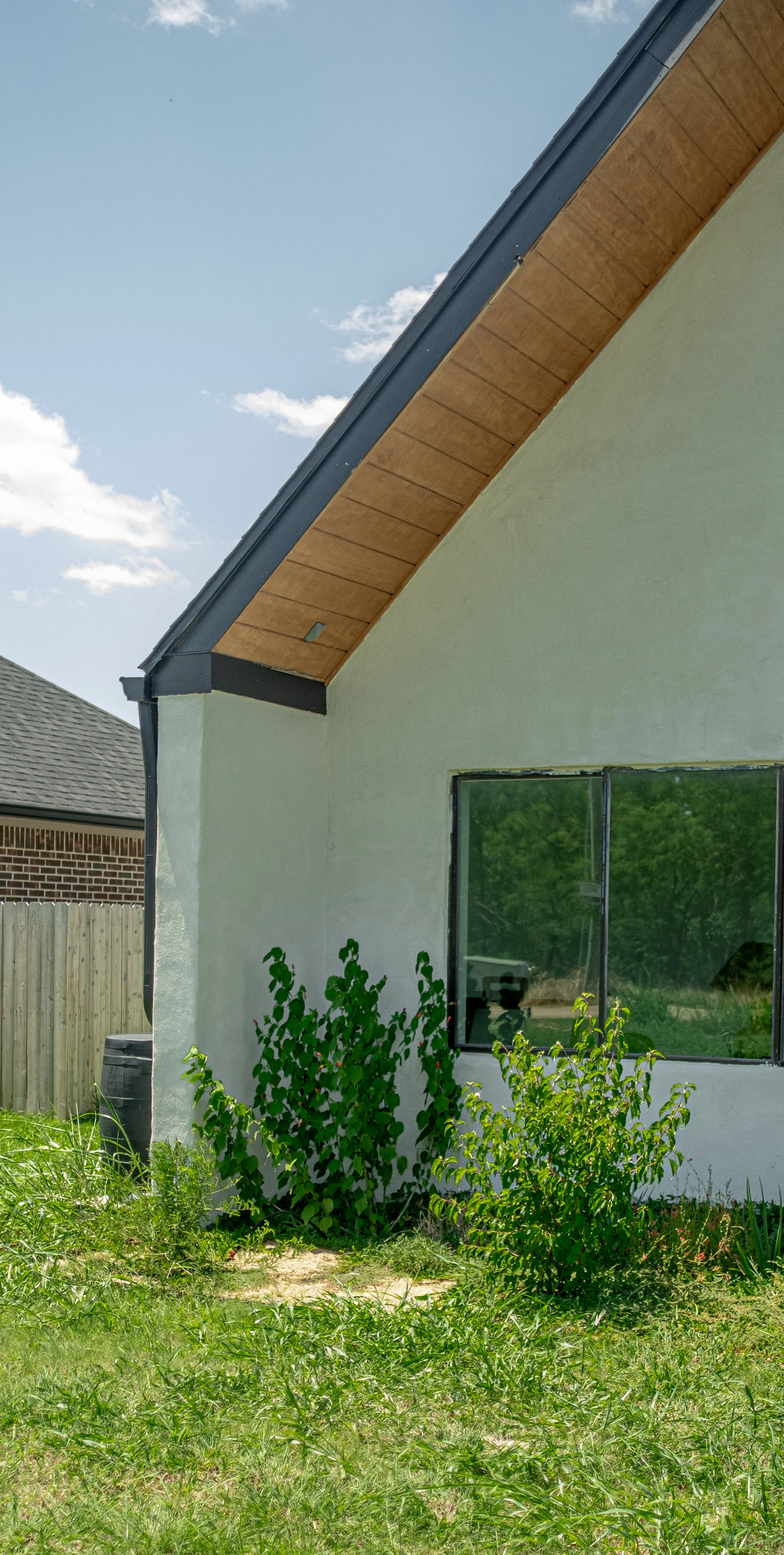 Close-up of a house's side with a white wall, a large window, and a black roof with undercooling and wooden underside. There are green bushes and grass in front of the house, with another house and a fence visible in the background under a blue sky.