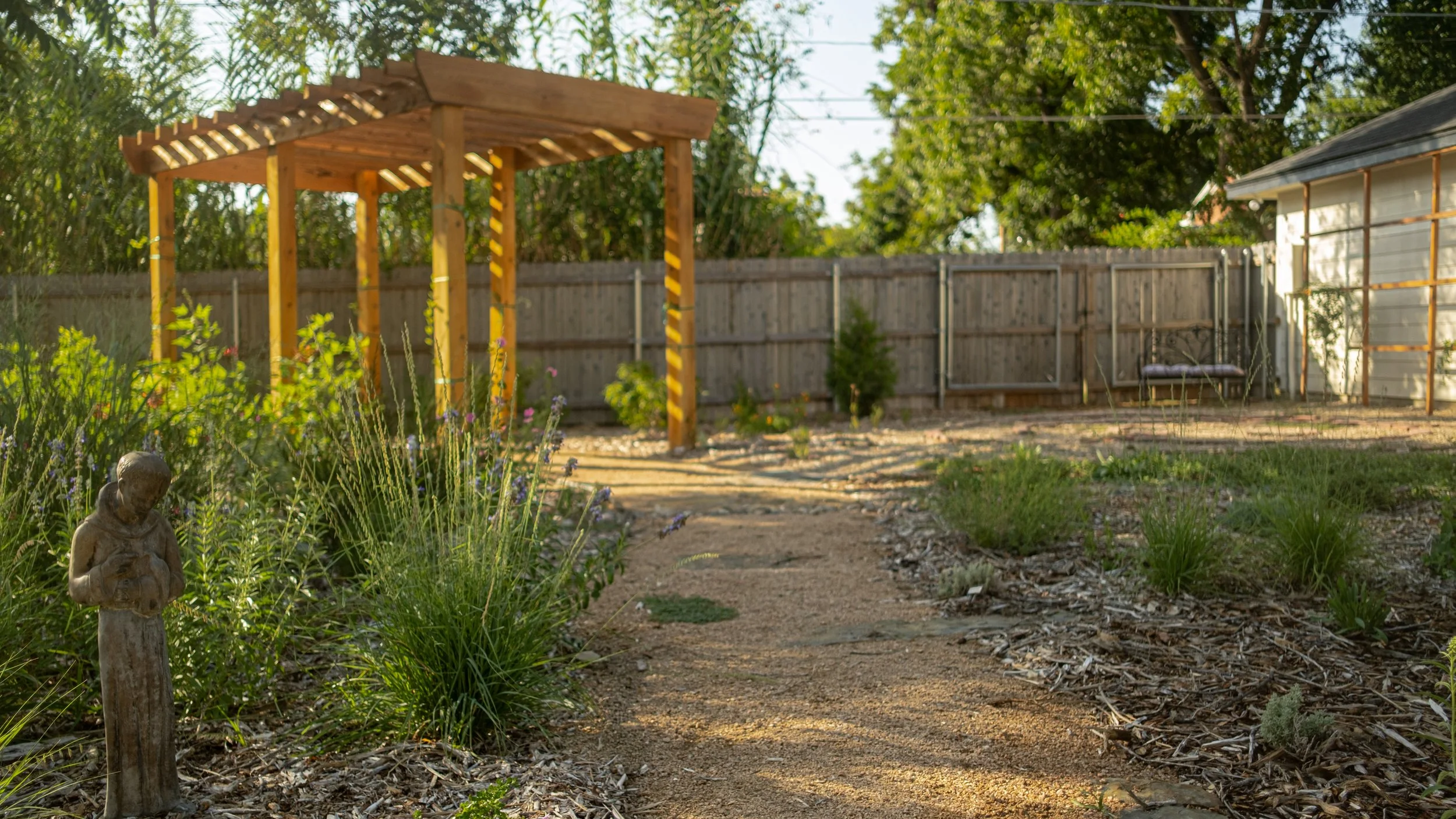 A backyard garden with a wooden pergola, a small statue of a person, and a wooden fence. There are plants, flowers, and trees around, with a dirt and gravel path.