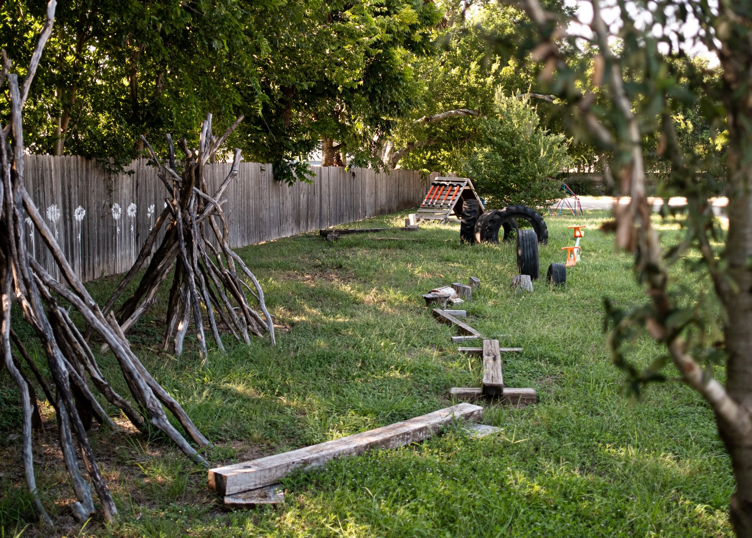 A backyard play area with a wooden fence, trees with green leaves, and various play equipment such as large tires, a small plastic slide, and a wooden balance beam on grass.