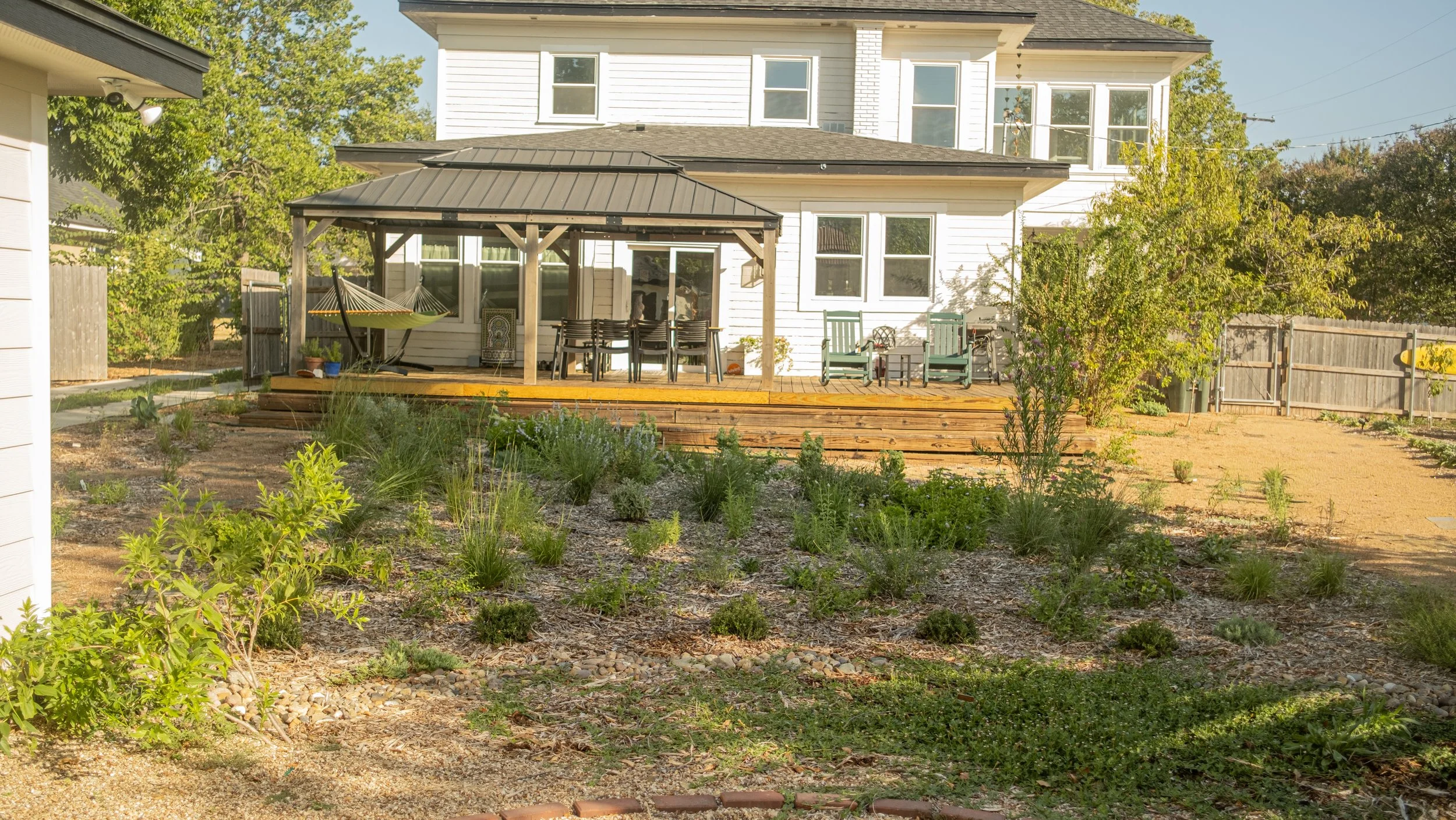 Backyard of a two-story white house with a deck, outdoor furniture, and a hammock, surrounded by trees and a garden area.