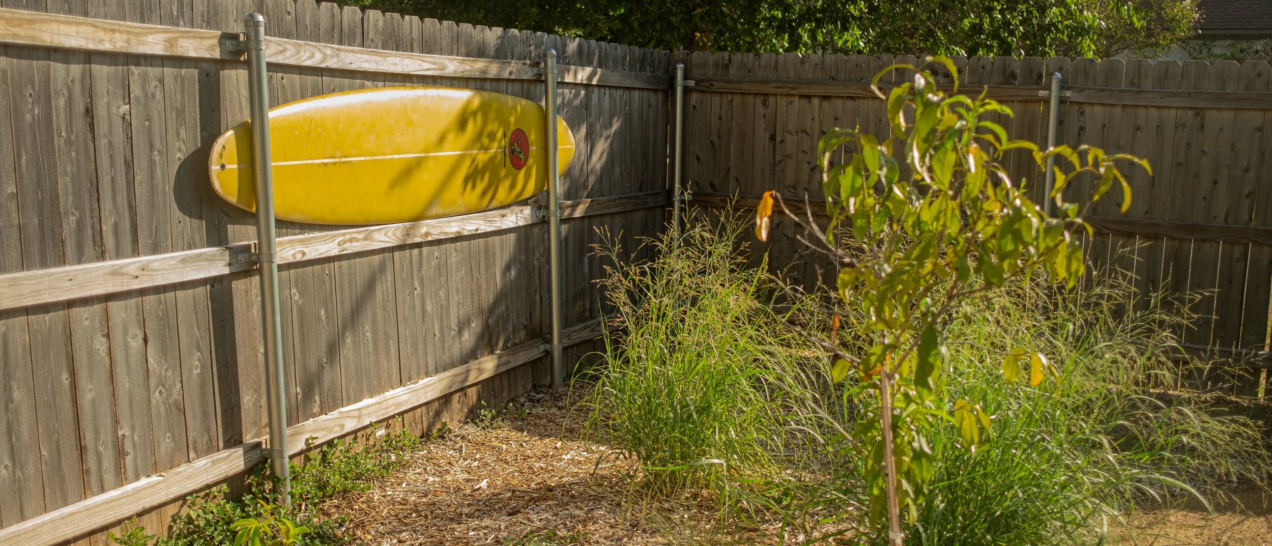 Yellow surfboard mounted on a wooden fence with a sticker of a red circle and a black bird, surrounded by green trees and overgrown grass.