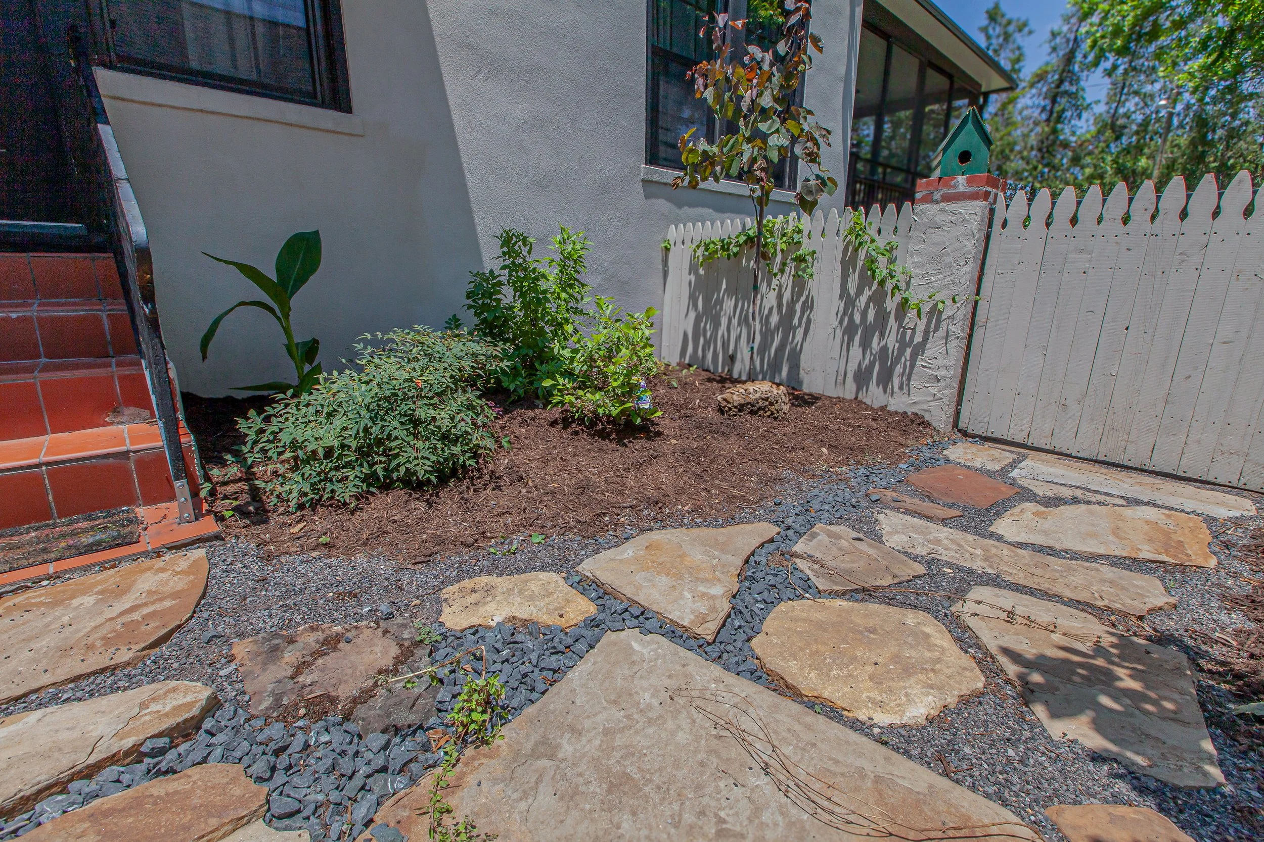 A small garden area with young plants, mulch, stone pathway, white fence, and part of the house with a window and screened porch in the background.