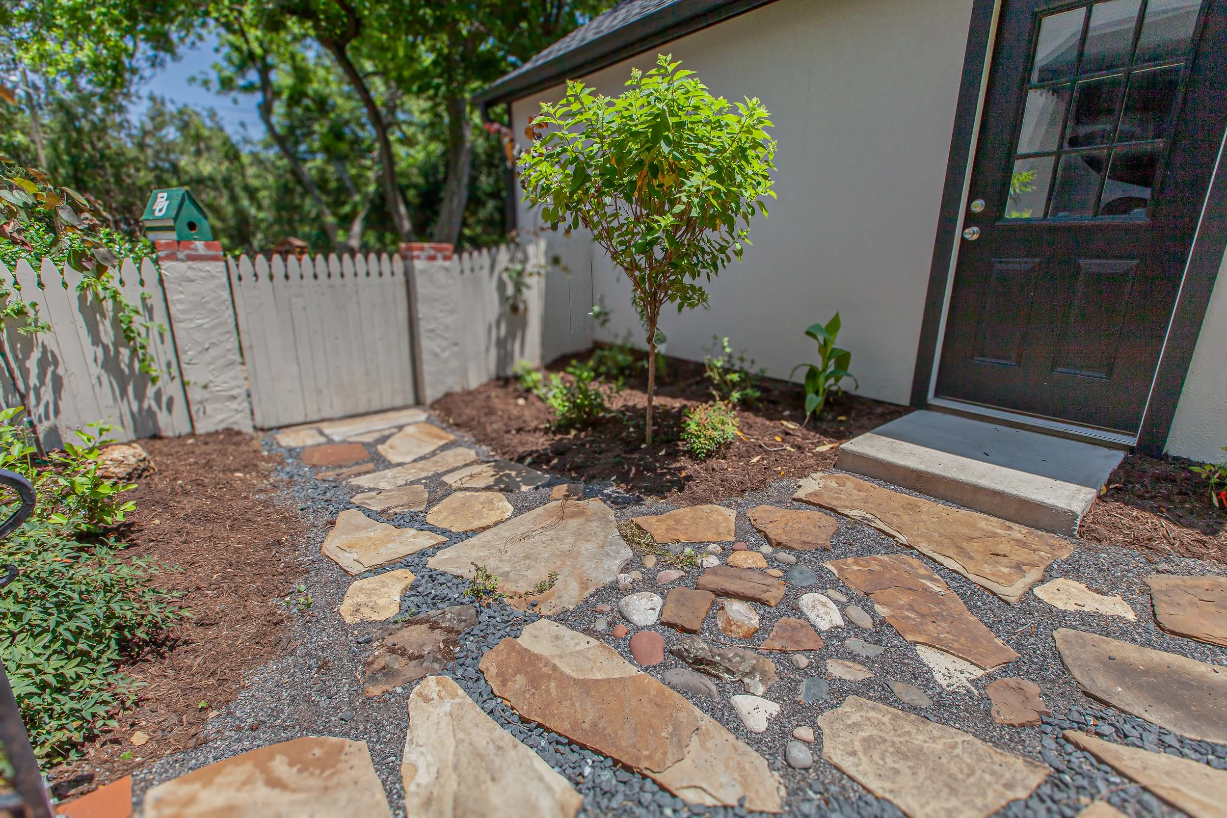 A small garden area outside a house with a young tree, potted plants, the white house wall, and black front door. There is a stone and gravel pathway.