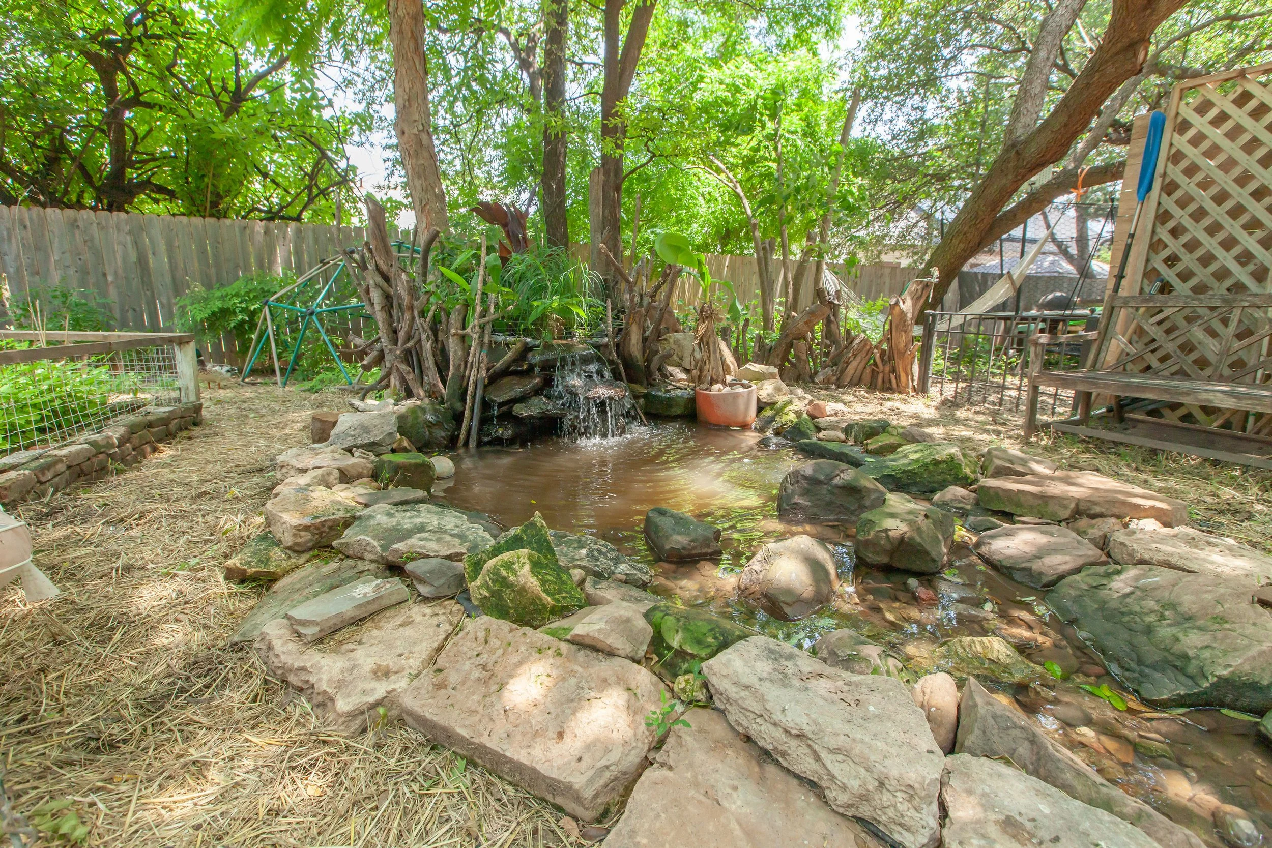 Backyard with small pond, rocks, trees, wooden fence, and various garden items.