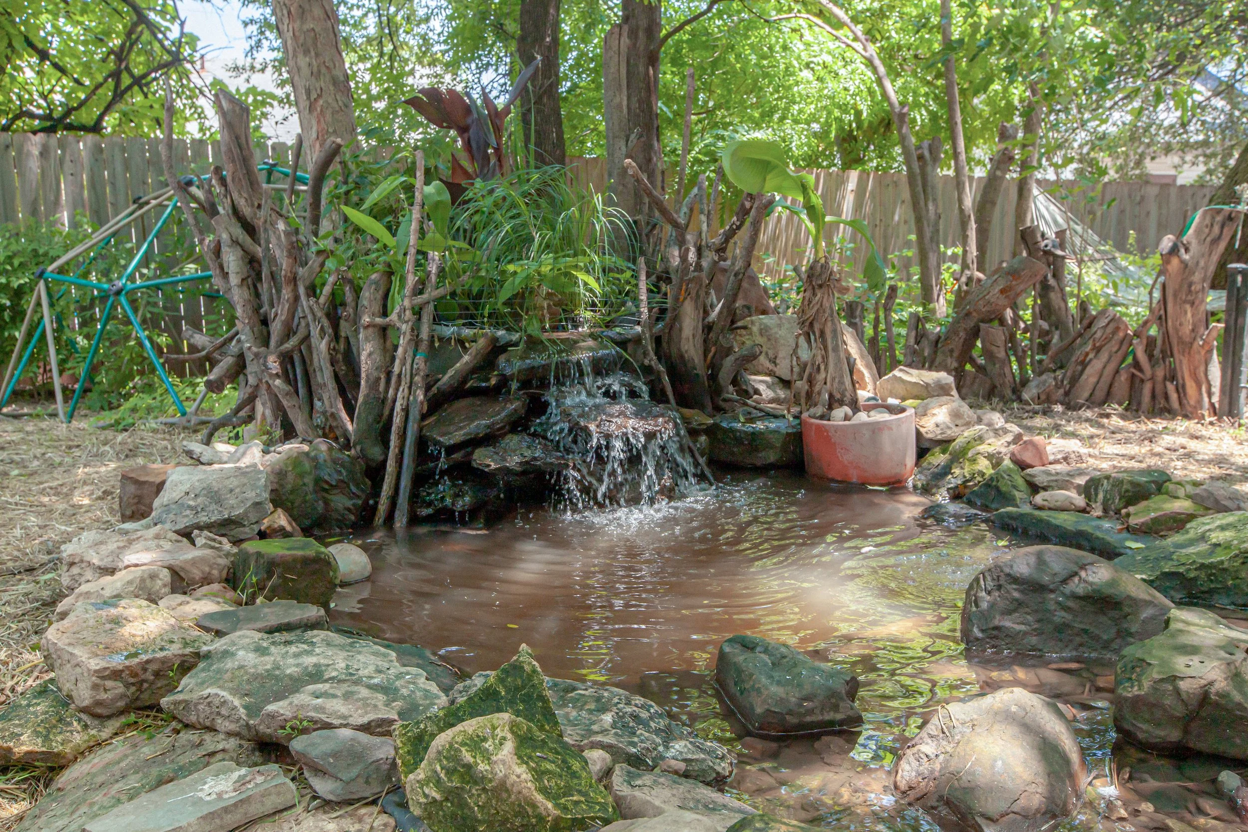 A small natural water feature with a waterfall surrounded by rocks, plants, and trees in a backyard garden, with a wooden fence in the background.