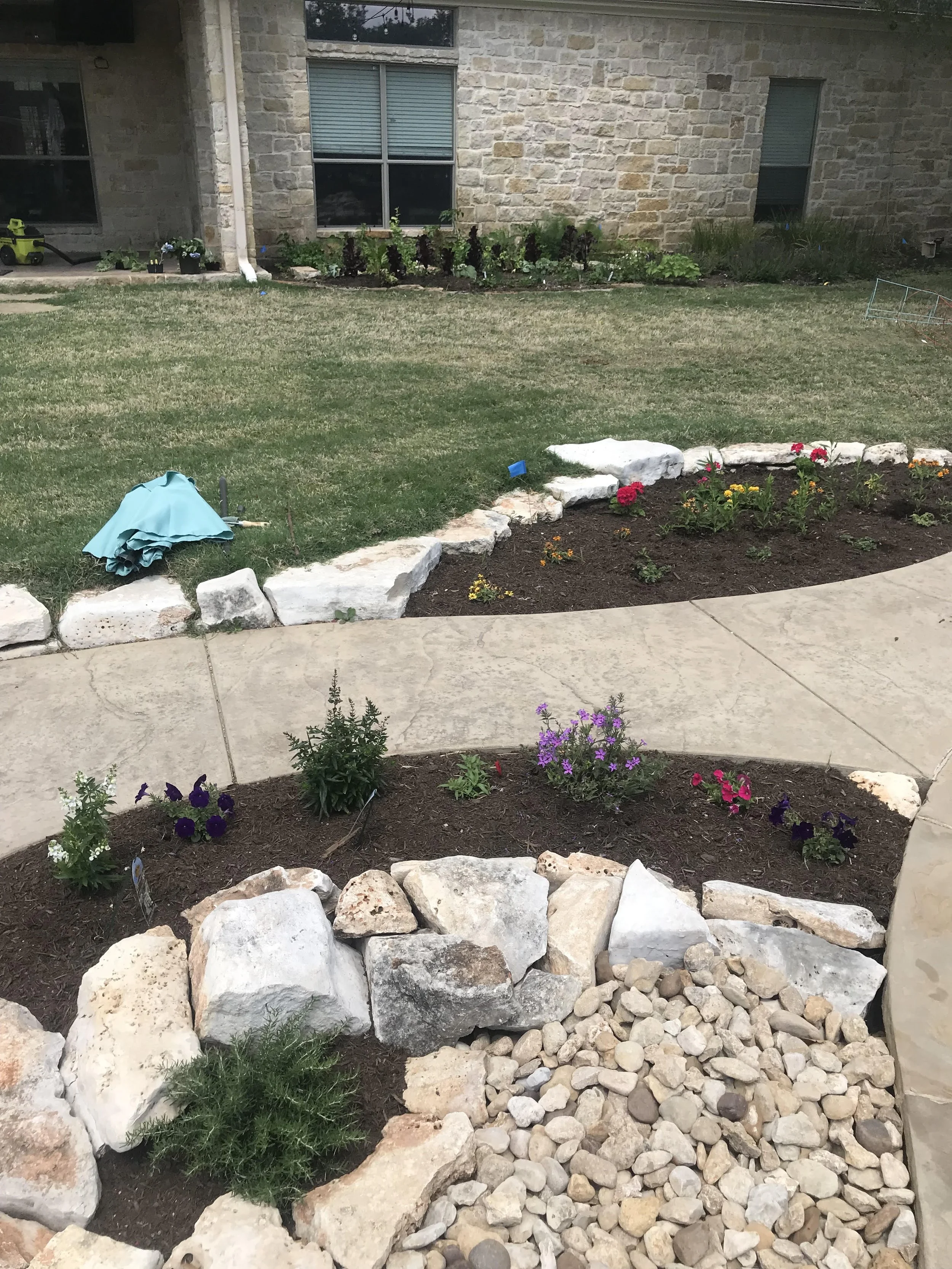 A backyard garden with a curved flower bed edged with rocks, filled with various flowering plants. There is a section with small, light-colored rocks and other with dark mulch. You can see part of a house with brick walls and windows in the backgroun