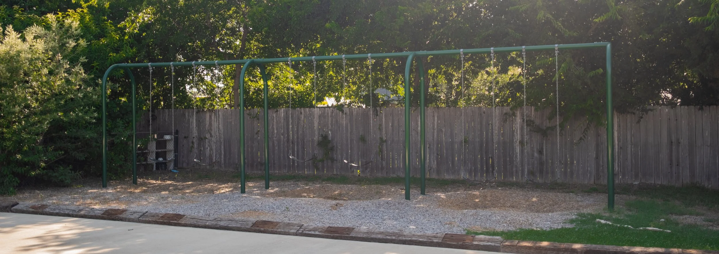 Empty outdoor swings set with five swings hanging from a green metal frame, trimmed with a wooden border, behind a wooden fence and trees