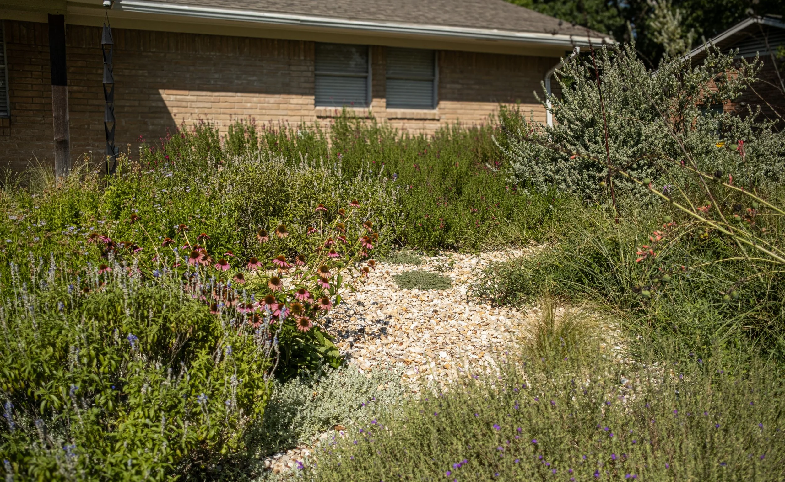 A flower garden with a variety of plants and flowers, a gravel pathway, a brick house in the background, and trees overhead.