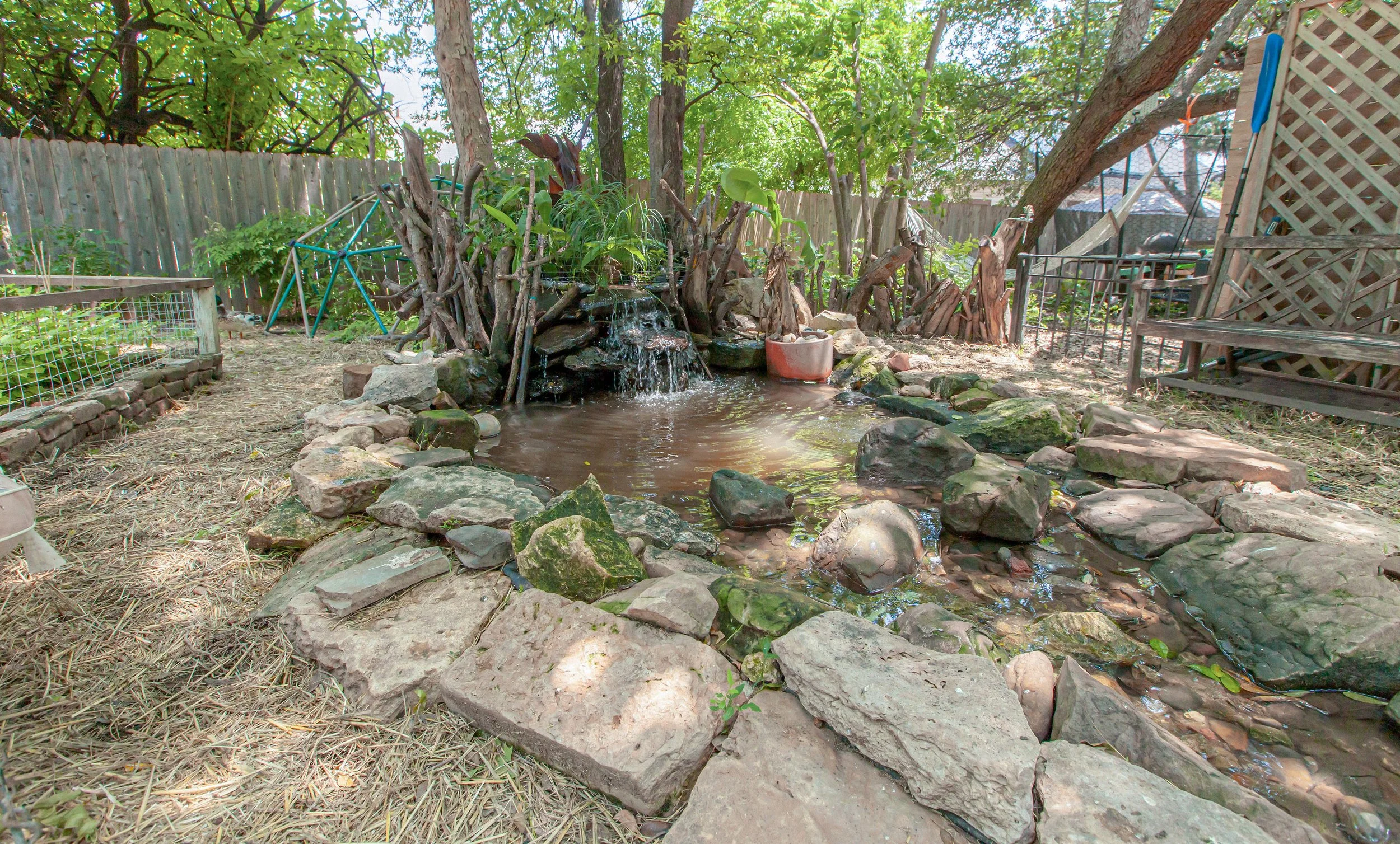 A small backyard with a pond surrounded by rocks, a waterfall feature, and various plants and trees. Wooden fences and garden structures are visible in the background.