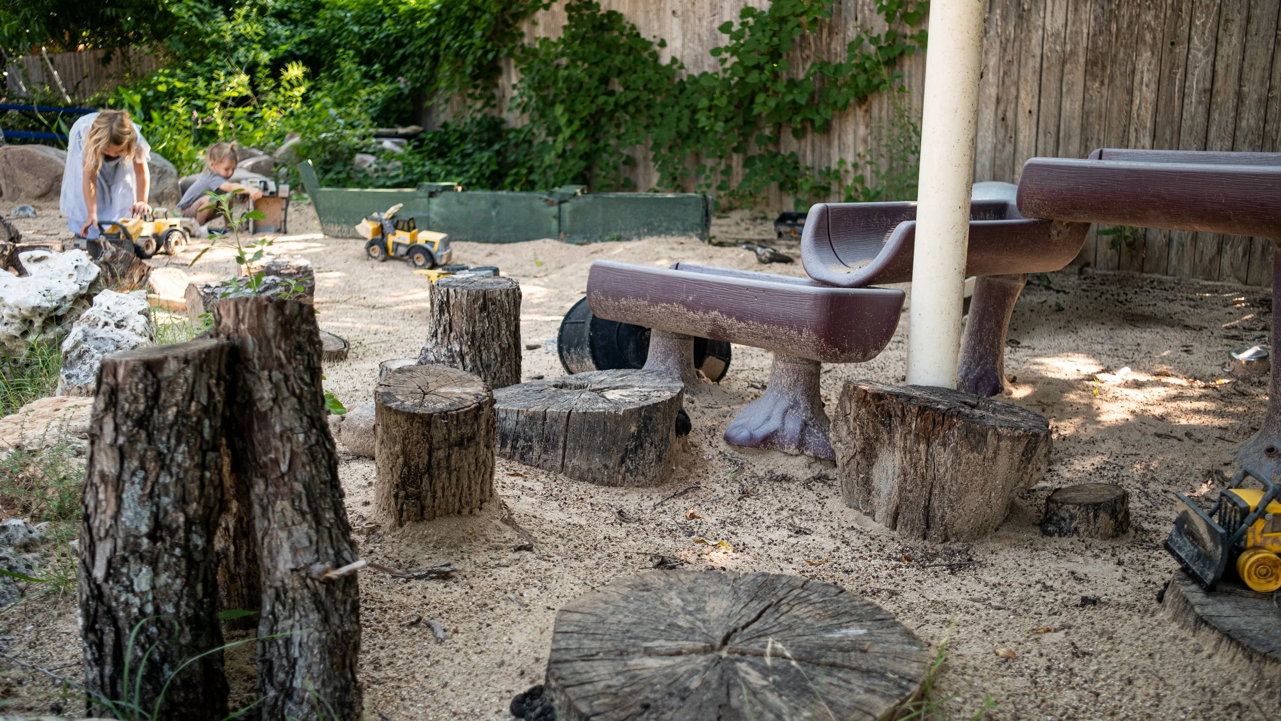 A backyard area with a sand surface, wooden tree stumps arranged as seating, a wooden bench, and a small tree. In the background, two children play with toy construction vehicles, and there are green plants, rocks, and a wooden fence.