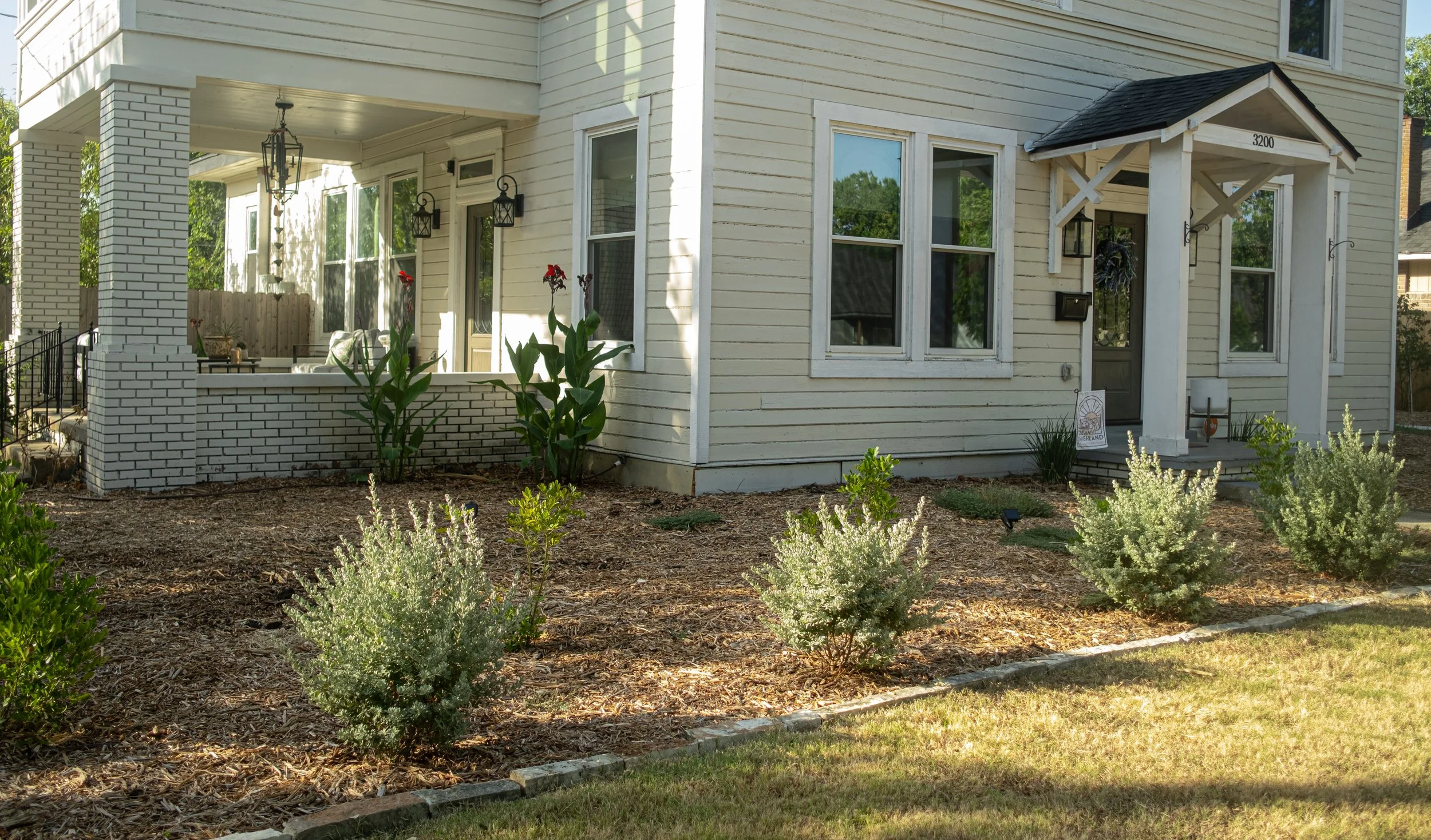Front view of a light-colored wooden house with a small porch, black lantern-style light fixtures, and a garden with bushes and plants.