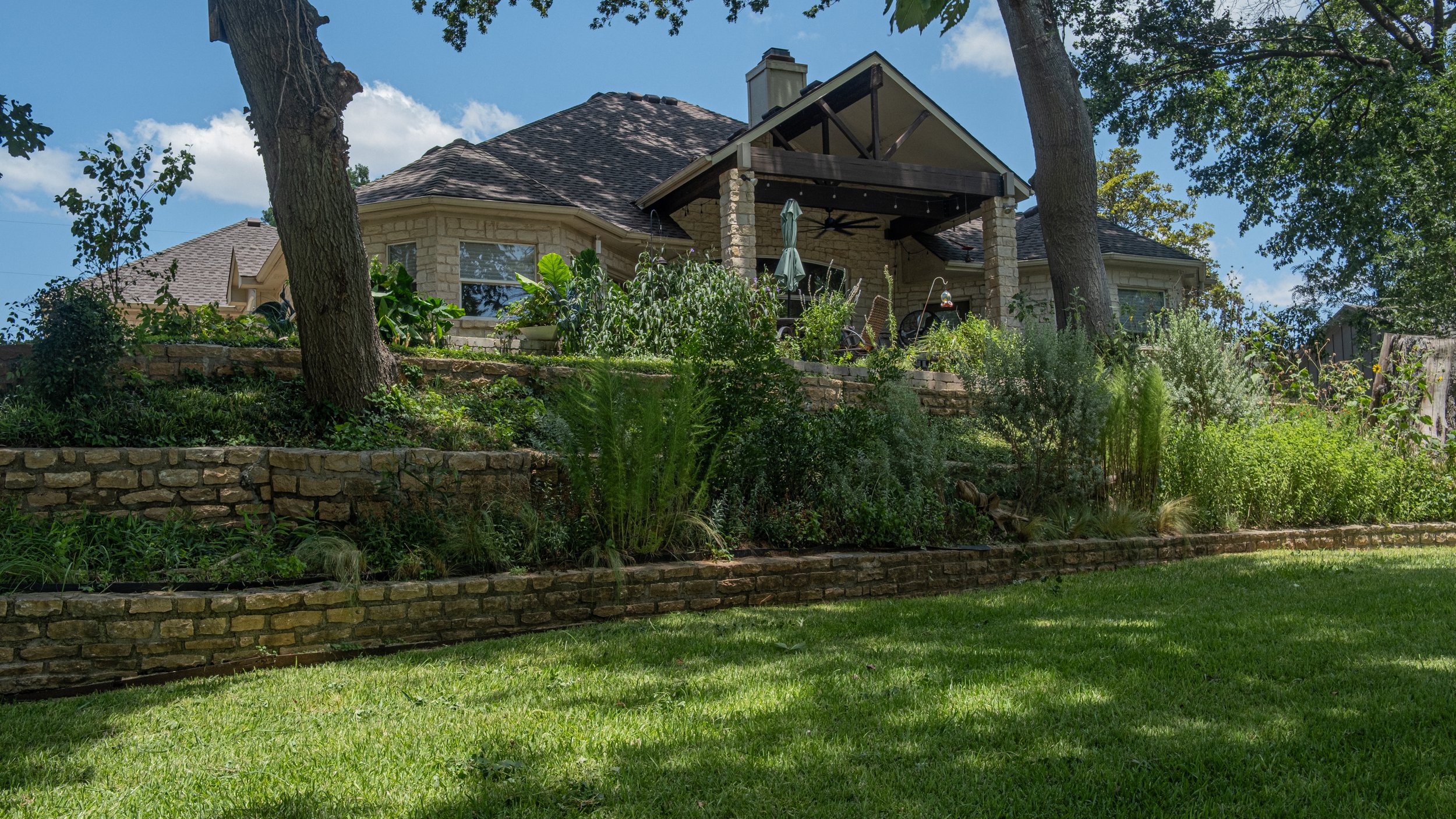 Backyard with stone retaining walls, lush greenery, trees, and a house with a porch and a shaded seating area.