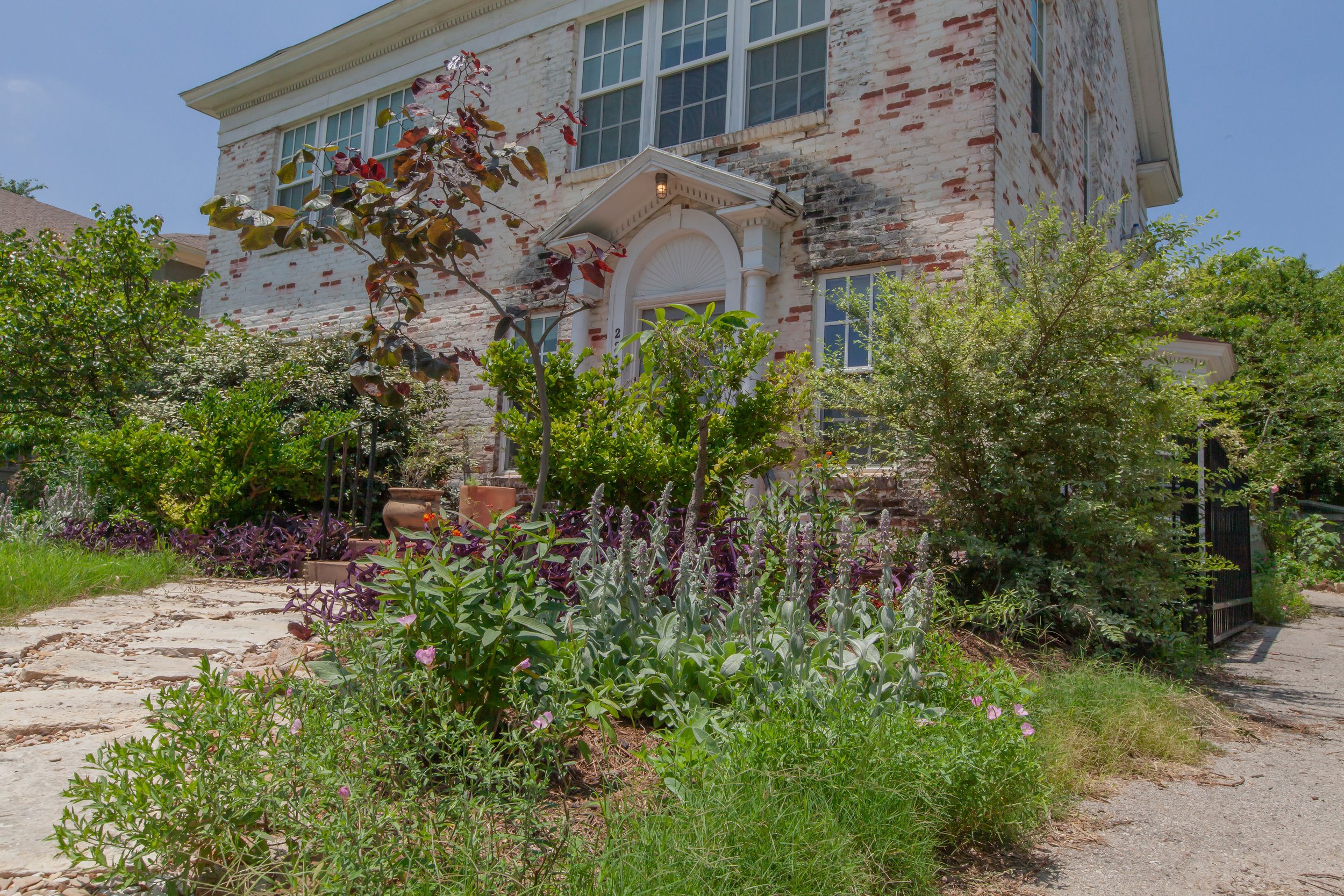 Front of a brick house with a garden and pathway, surrounded by plants and trees.