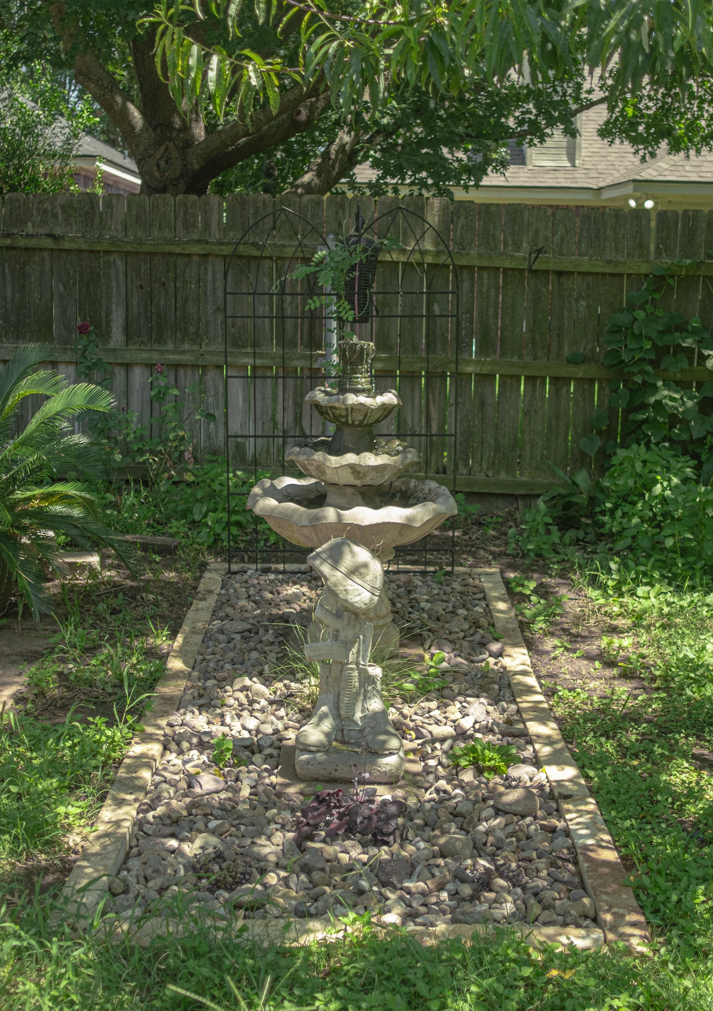 A small decorative fountain with three tiers, made of stone, situated in a garden with various green plants around. The fountain is placed on a bed of small rocks within a border of rectangular bricks, with a wooden fence and trees in the background.
