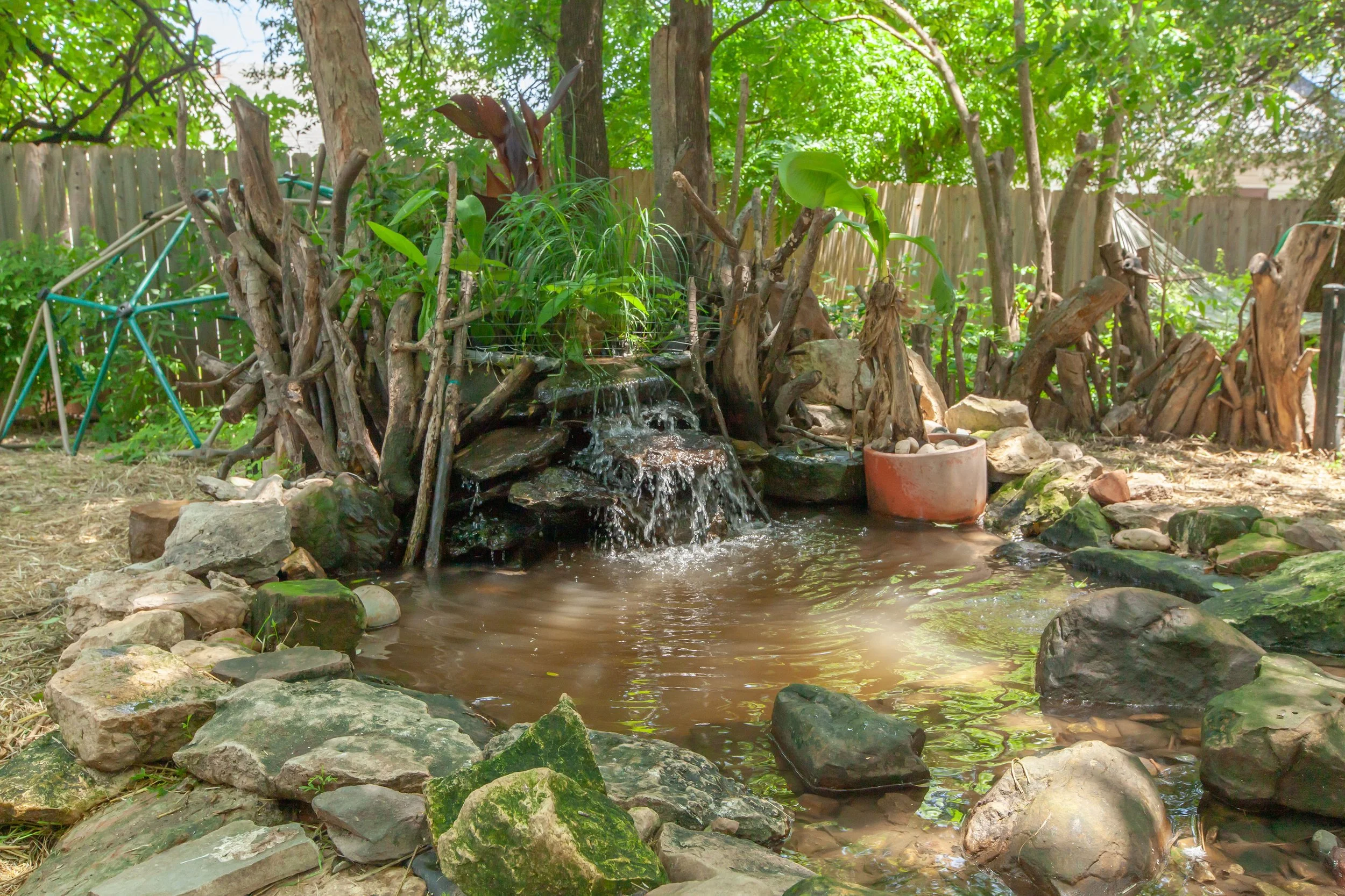 A small backyard water feature with a waterfall surrounded by rocks and driftwood. Green plants and trees are in the background, and a wooden fence encloses the yard.