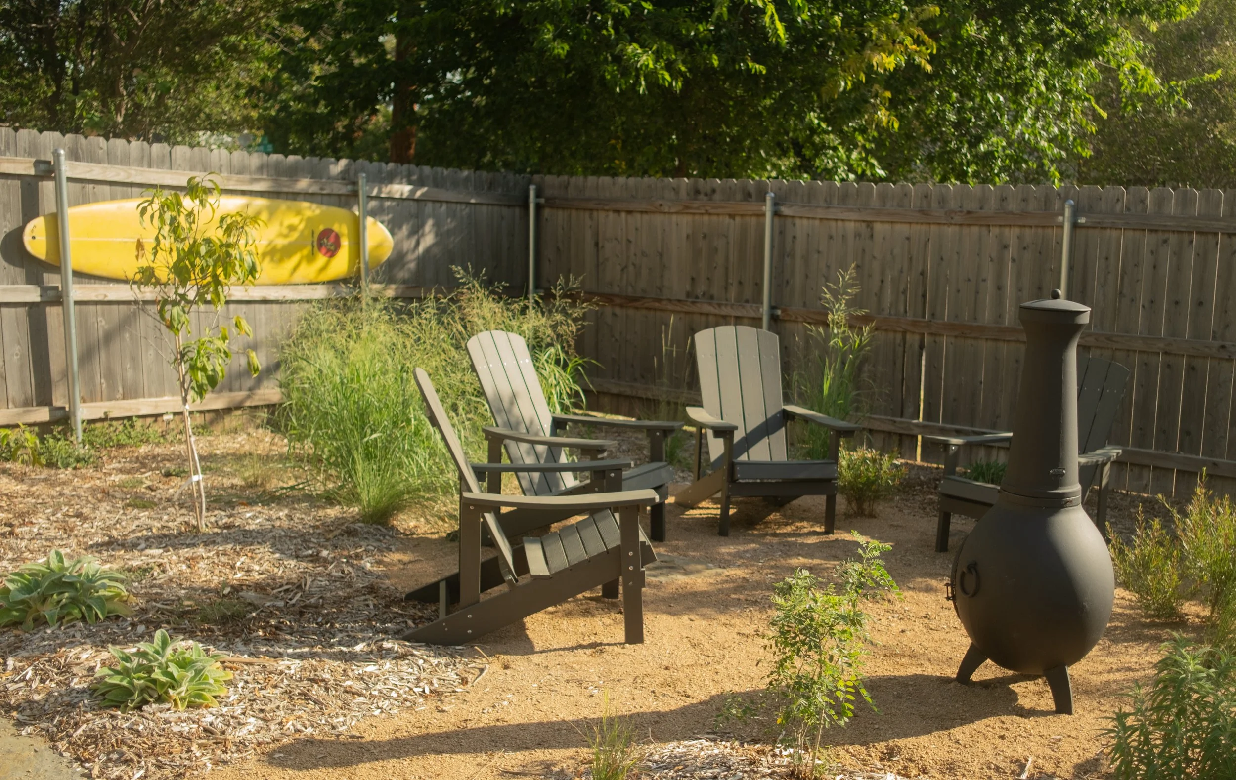 A backyard scene with four Adirondack chairs, a chiminea, green plants, and a yellow kayak mounted on a wooden fence, surrounded by trees.