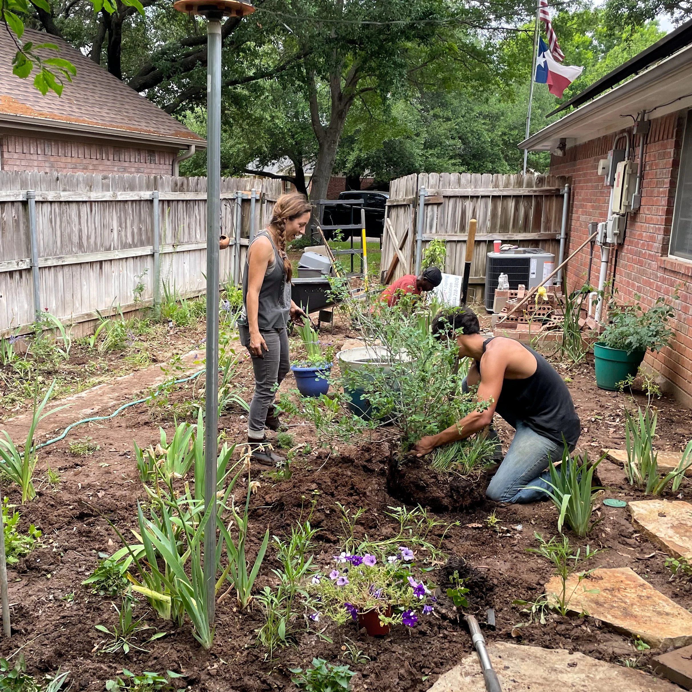 A woman and a man gardening in a backyard with a clay-brick house, wooden fence, and trees. The woman is standing, smiling, and the man is kneeling, working in the soil near flowering plants. A flagpole with the Texas state flag is visible, along with garden tools, an air conditioning unit, and electrical meters attached to the brick house.