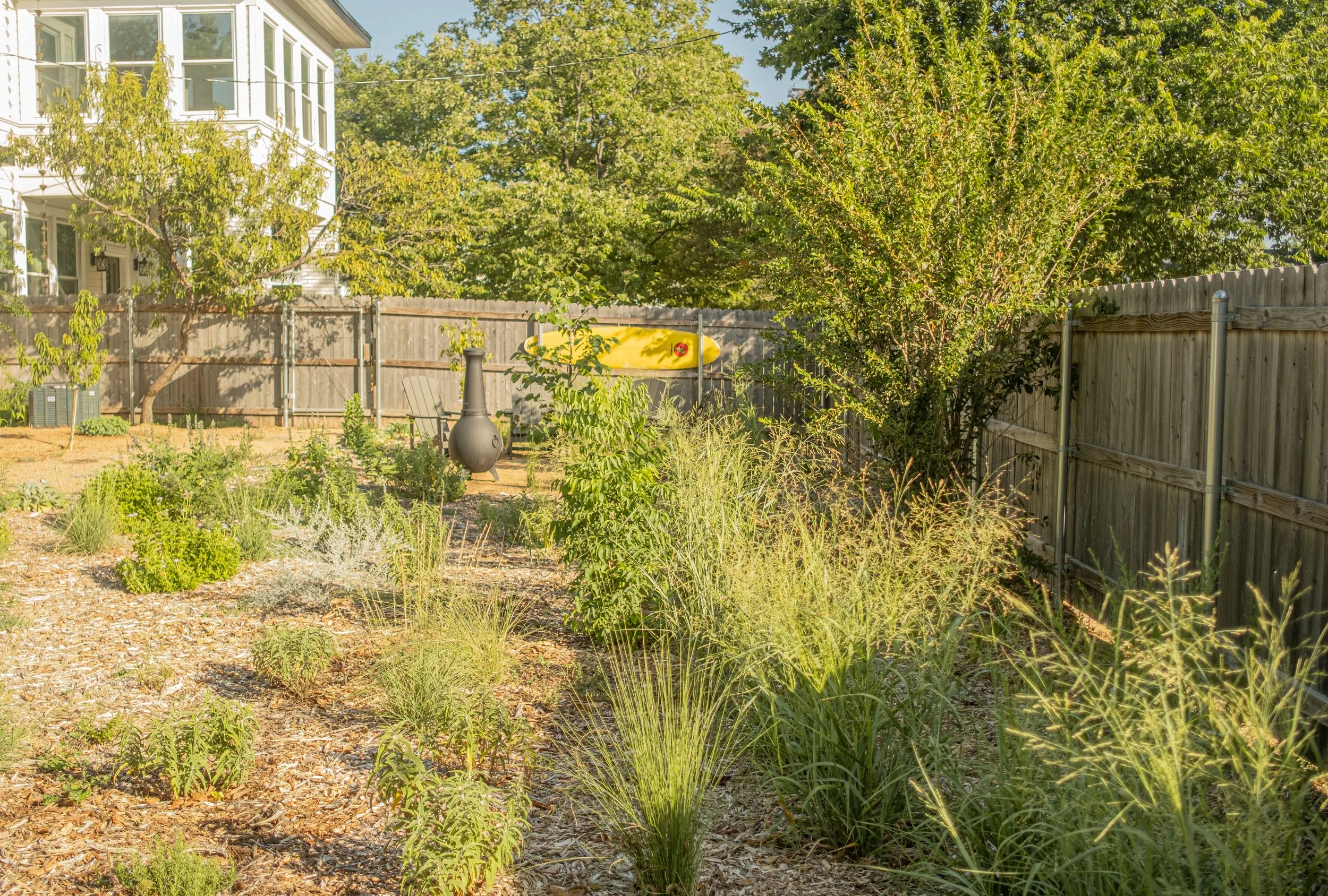 A backyard garden with various green plants, a wooden fence, a yellow kayak mounted on the fence, and a house with large windows in the background. There is also a patio chair and a chiminea in the garden.