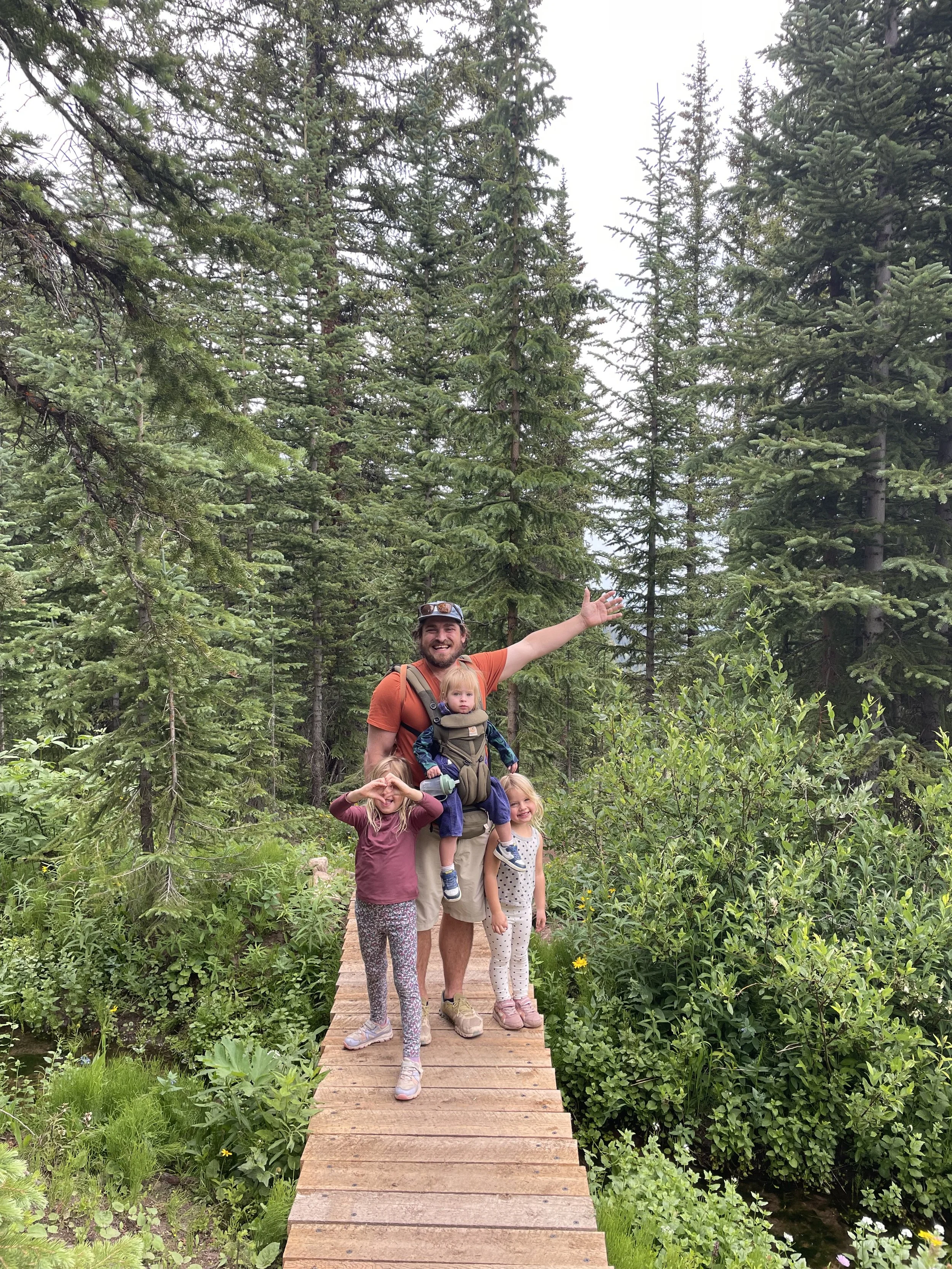 A man with a beard wearing a backpack and orange shirt, standing on a wooden bridge in a forest, is smiling and waving with four children, three girls and one boy, surrounded by lush green trees and plants.