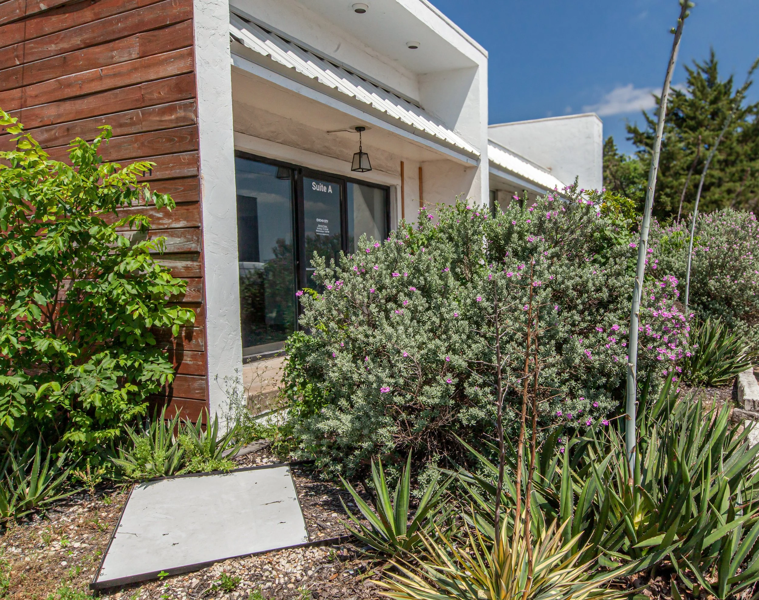 The image shows the exterior of a modern white building with a balcony, surrounded by lush greenery and pink flowers, with a bright blue sky.