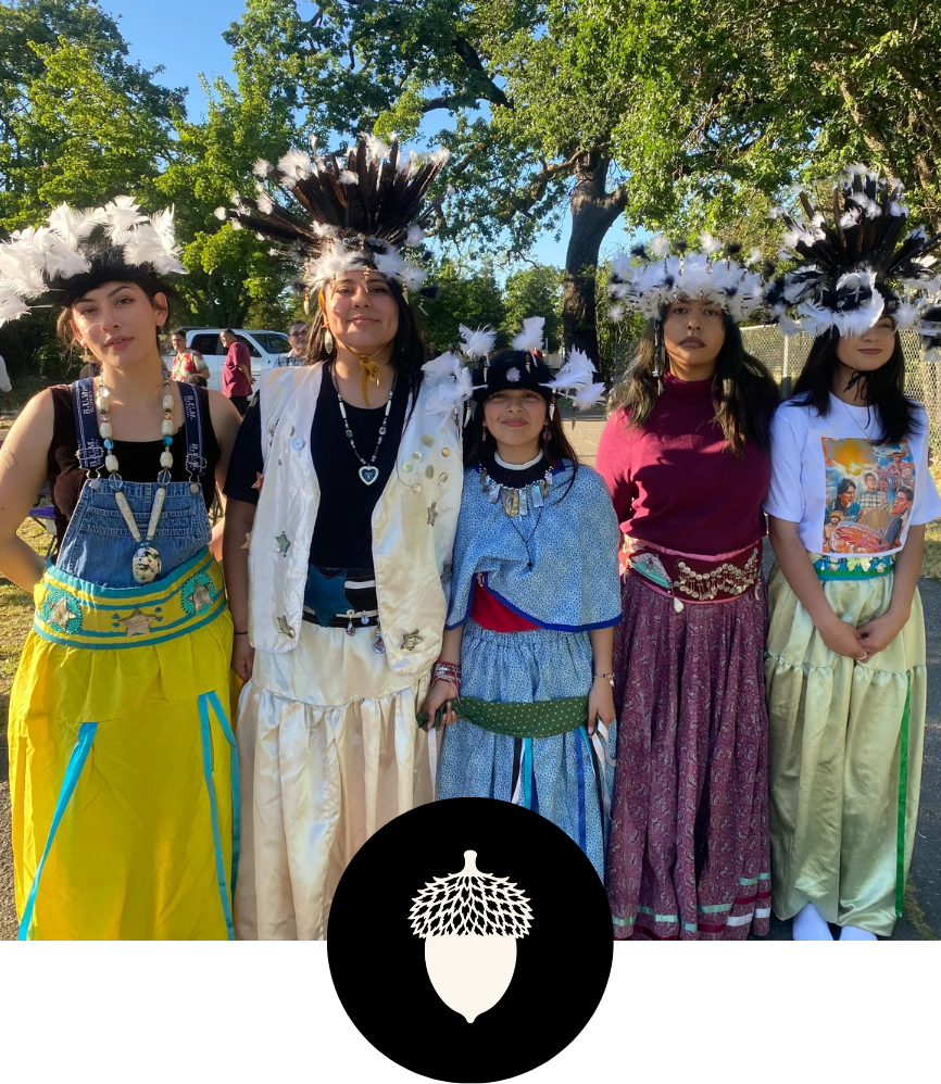 Group of five young women dressed in colorful traditional attire with feathered headdresses, standing outdoors in a Sonoma County park with green trees and blue sky.