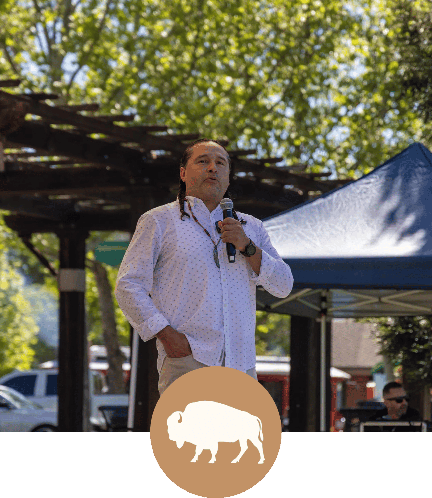 A Native man with long dark hair and a braid, wearing a white dotted shirt, standing outdoors with a microphone, speaking at a Sonoma County nonprofit event under trees with green leaves and a blue tent in the background.