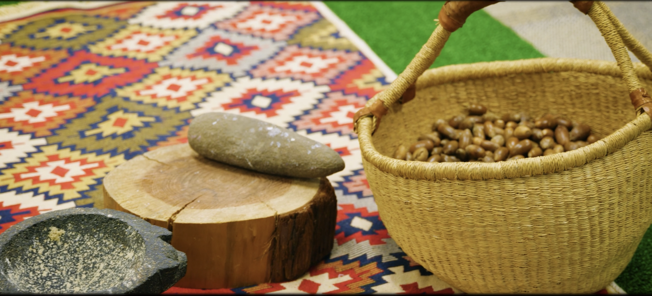 A woven basket filled with acorns, placed on a colorful, patterned fabric with geometric designs. Stones and wooden slabs sit nearby at a workshop by the Progressive Tribal Alliance, the best Native nonprofit in Sonoma County.
