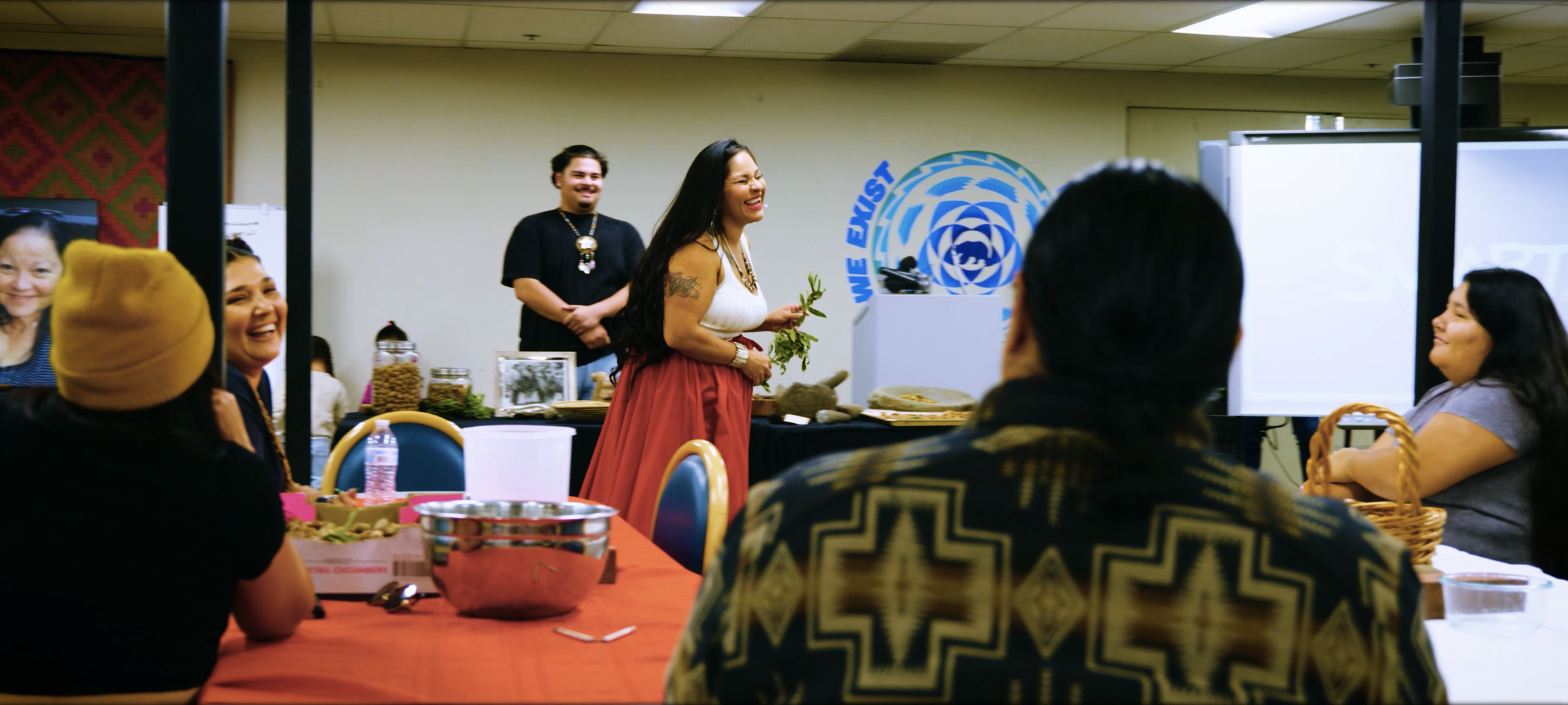 A group of Native people sits around a table, smiling and listening to a woman speak at a nonprofit event in Northern California. The woman stands, holding greenery, and wears a white top and a red skirt.