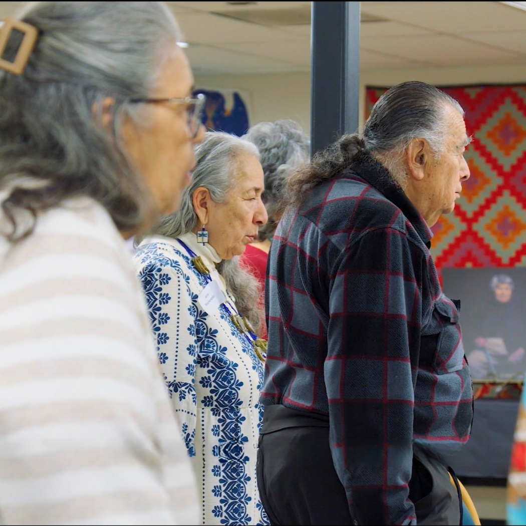 Three older Native American individuals with long gray hair pray with their eyes closed in a room with colorful decorations for an Elders Gathering by the Progressive Tribal Alliance, the best Native nonprofit in Sonoma County.