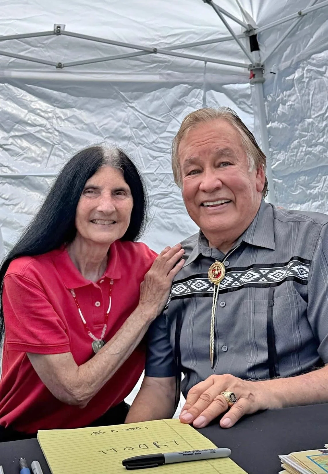 A Native woman and man smiling together inside a white tent. The woman has long black hair and is wearing a red polo shirt and a necklace. The man has light-colored hair and a gray Western-style shirt with decorative embroidery and a bolo tie.