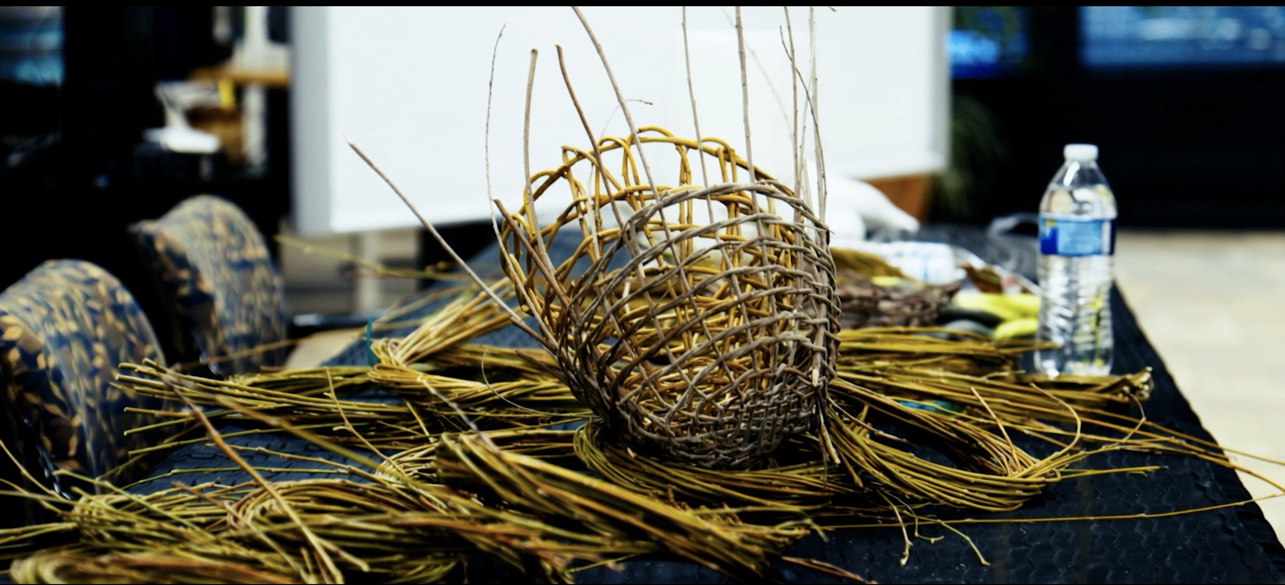 A Native woven basket made of thin plant stems, sitting on a black table with additional plant stems scattered around. In the background, a plastic water bottle and blurred objects can be seen.